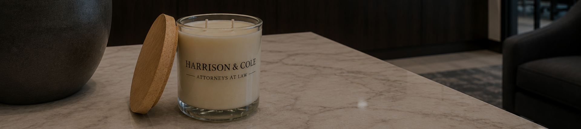 A white candle with a wooden lid on a marble table, with a large dark pot on the left and a black sofa and windows in the background.