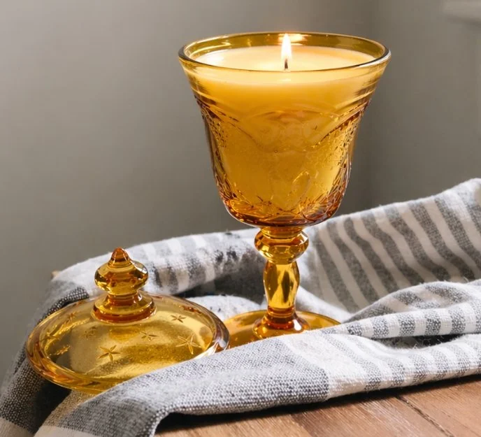 A lit yellow candle in a decorative glass holder and a matching glass goblet on a white and gray striped cloth on a wooden surface.