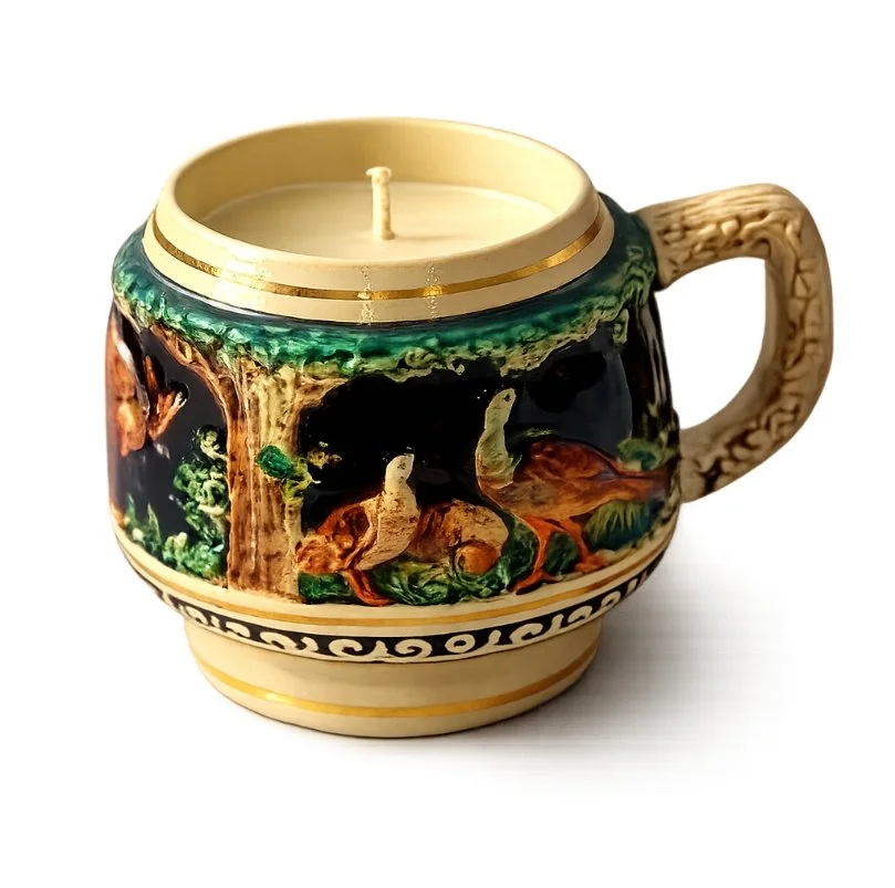Ceramic mug with a carved handle, decorated with a forest scene featuring trees, rocks, and moss, used as a candle holder with an unlit candle inside.