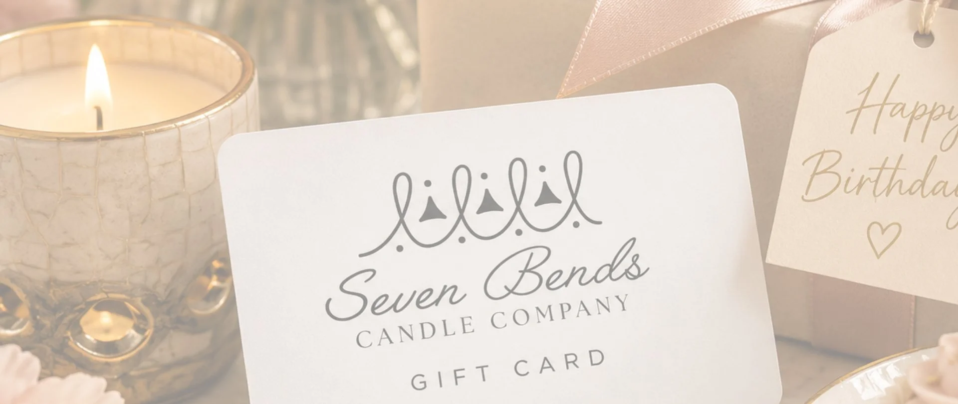 A lit candle in a textured silver holder, birthday gift card from Seven Bends Candle Company, and a 'Happy Birthday' card with gold lettering and a heart, on a light-colored surface.