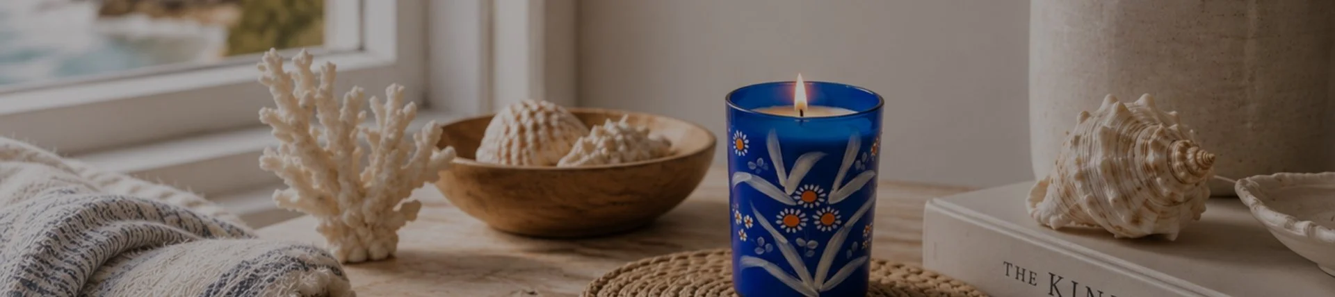 A coastal-themed table decor scene with a lit blue candle, seashells, coral, a wooden bowl with shells, and a large shell on a stack of books near a window.