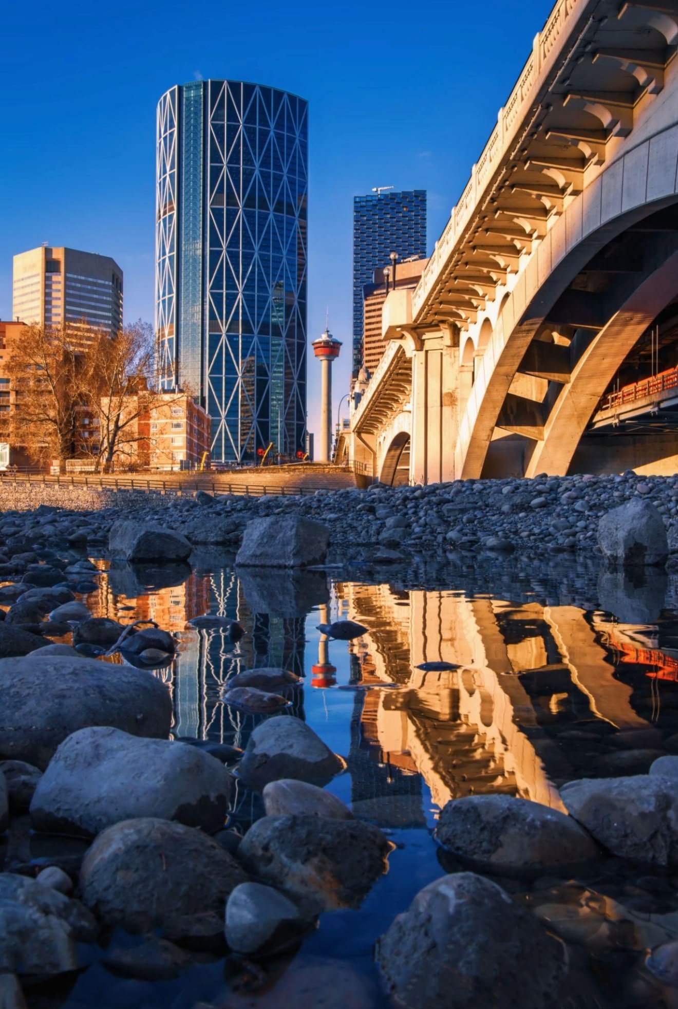 City skyline with tall modern buildings, a bridge, and a tv tower, reflected in a rocky river with water and stones in the foreground, during late afternoon or early evening.