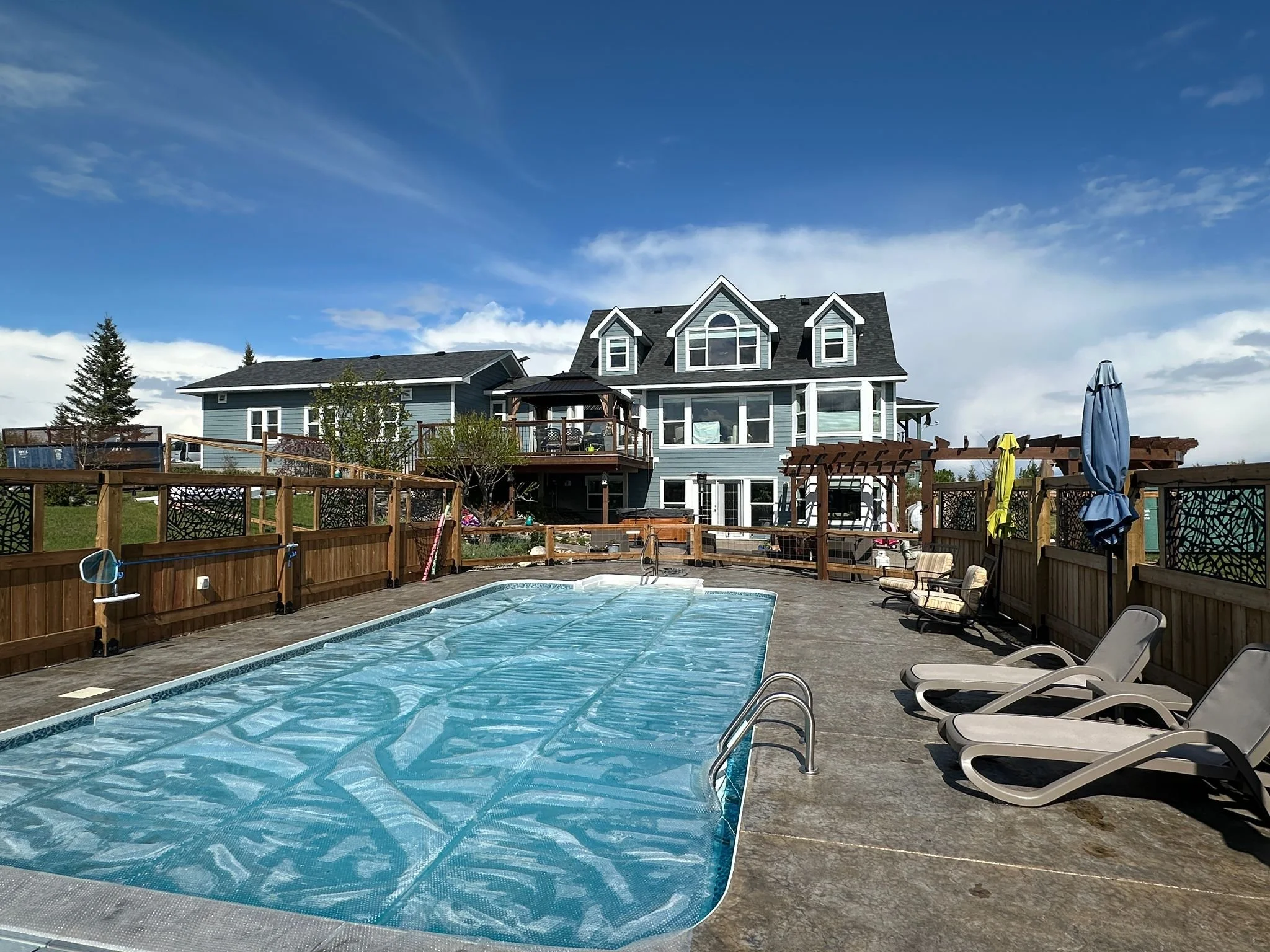 Backyard with swimming pool covered by a blue safety cover, surrounded by wooden fencing, with a multi-story house in the background under a partly cloudy sky.