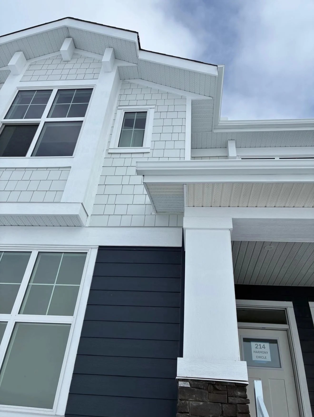 Close-up of a modern white and black house exterior with large windows, horizontal black siding, and white trim. The house has detailed eaves and a stone base by the front door, with a sign that reads '214 HARMONY CIRCLE'.