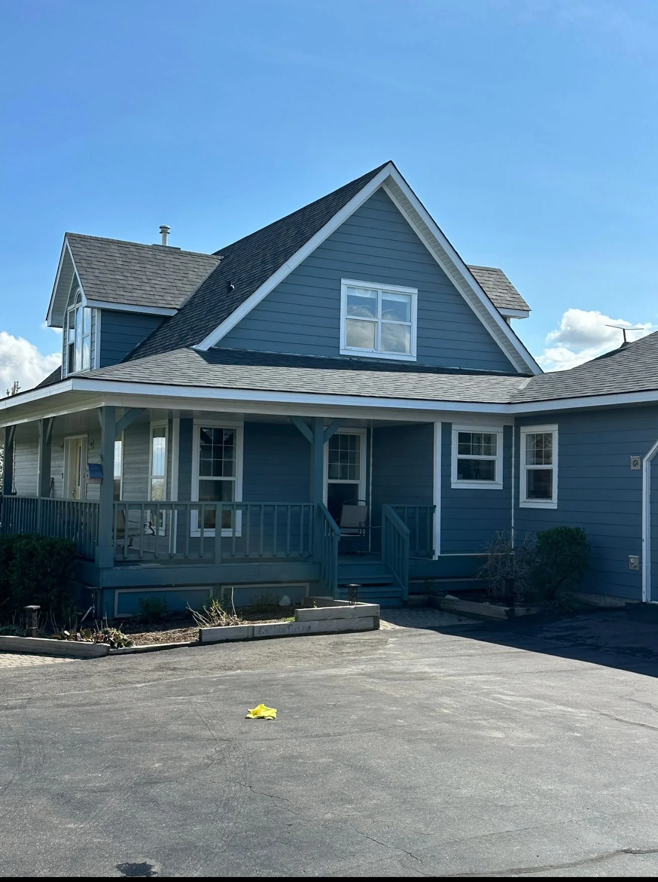 A blue two-story house with a covered porch, multiple windows, and a gabled roof, set against a partly cloudy sky. The front yard has a small garden bed and a paved driveway.