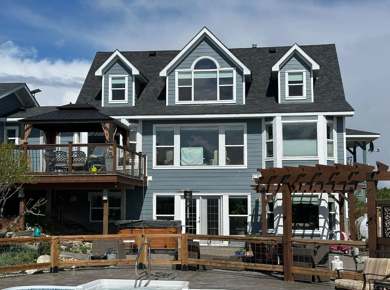 A large blue multi-story house with a ship shape roof, multiple windows, and a wooden deck with furniture and a hot tub in the backyard.