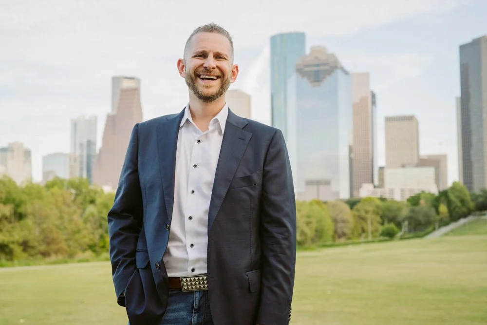 Dr. Ty Lerman smiling in a suit, standing outdoors with the Houston skyline and green park in the background.