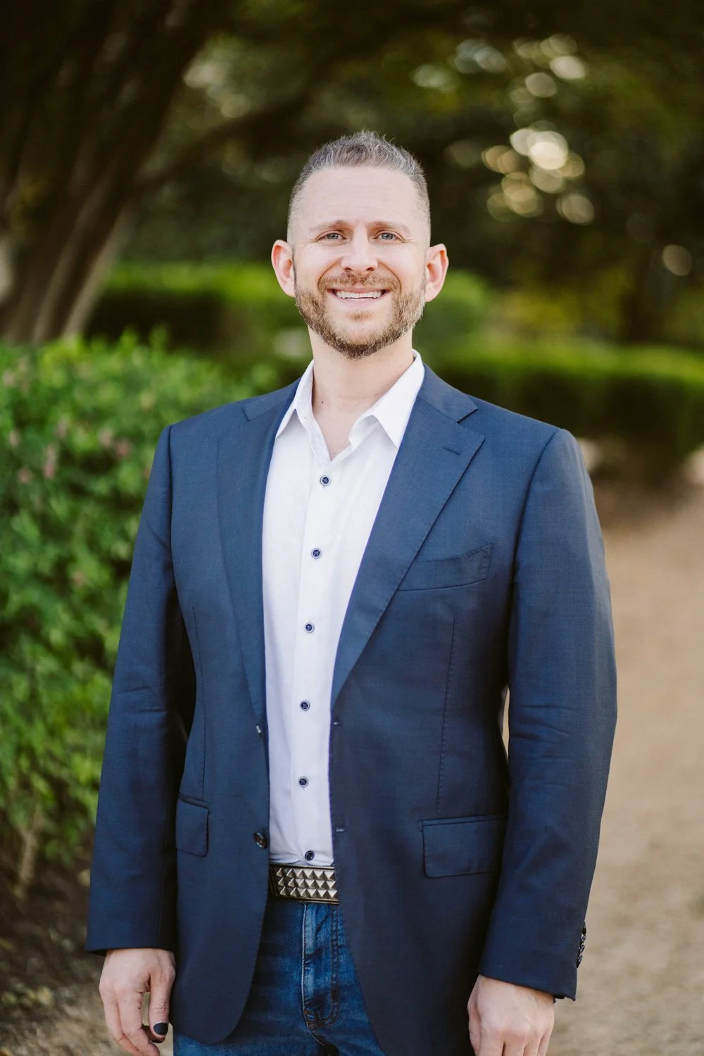 Dr. Ty Lerman is smiling in an outdoor setting with a blurred background of trees and greenery. He is dressed in a navy blue blazer, a white button-up shirt, and dark jeans.
