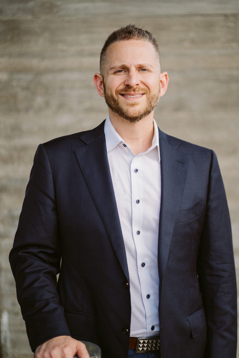 Dr. Ty Lerman smiling in a dark suit and white shirt standing in front of a textured grey wall.