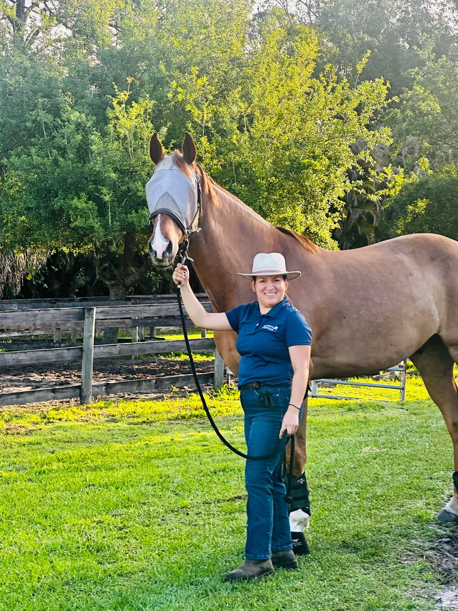 Mariana herself, standing on grass holding a tall horse's lead rope, with trees and a wooden fence in the background.