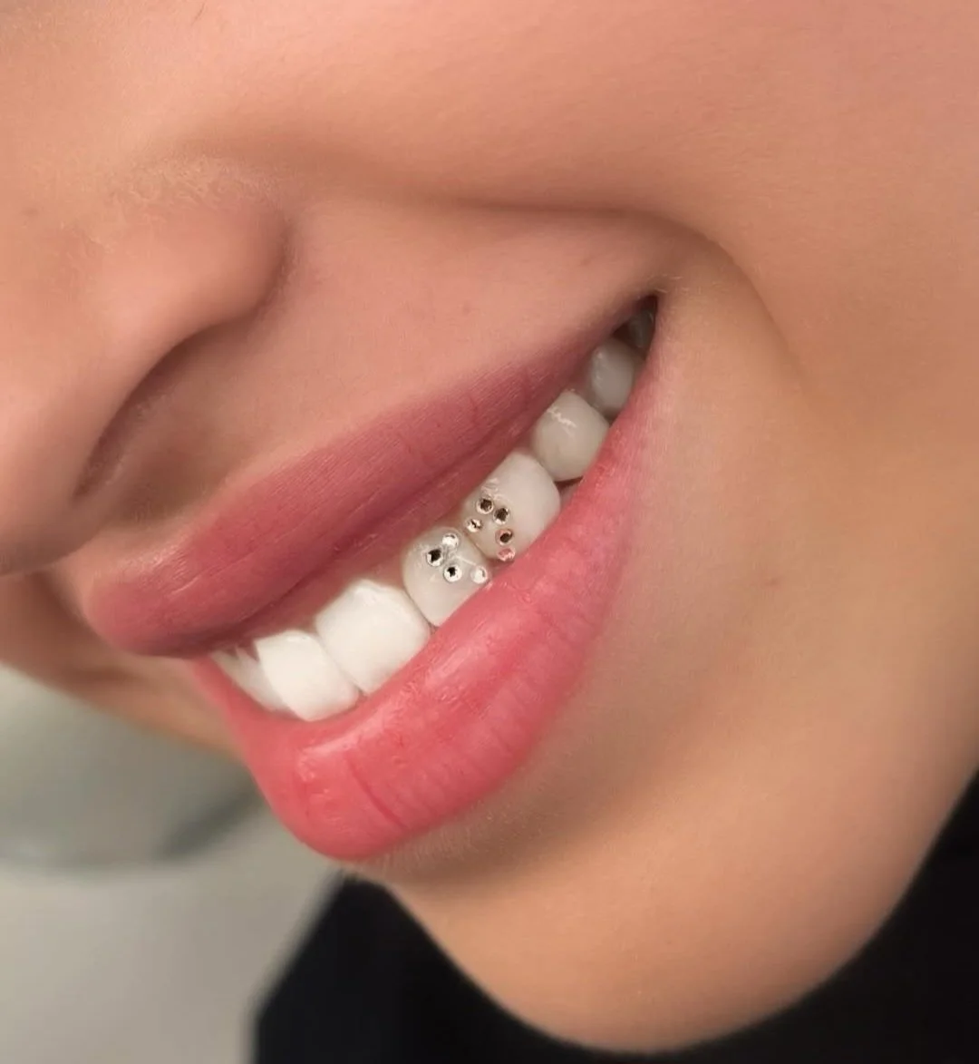 Close-up of a person's face showing a partial smile with white teeth and pink lips, with a small piercing on the lower lip.