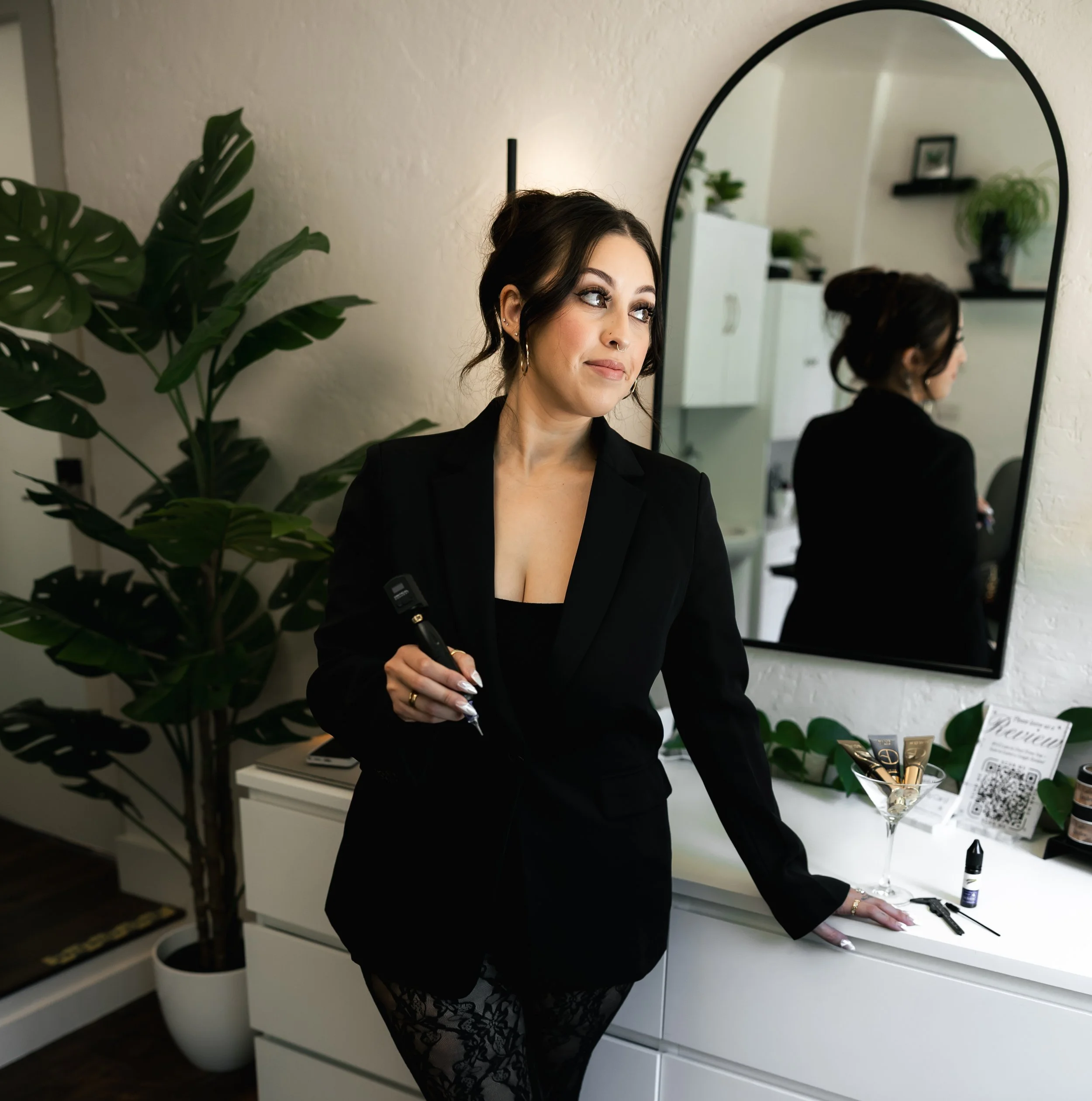 A woman with dark hair styled in an updo, wearing makeup and earrings, dressed in black lace clothing, stands in front of a mirror in a room decorated with plants and shelves.