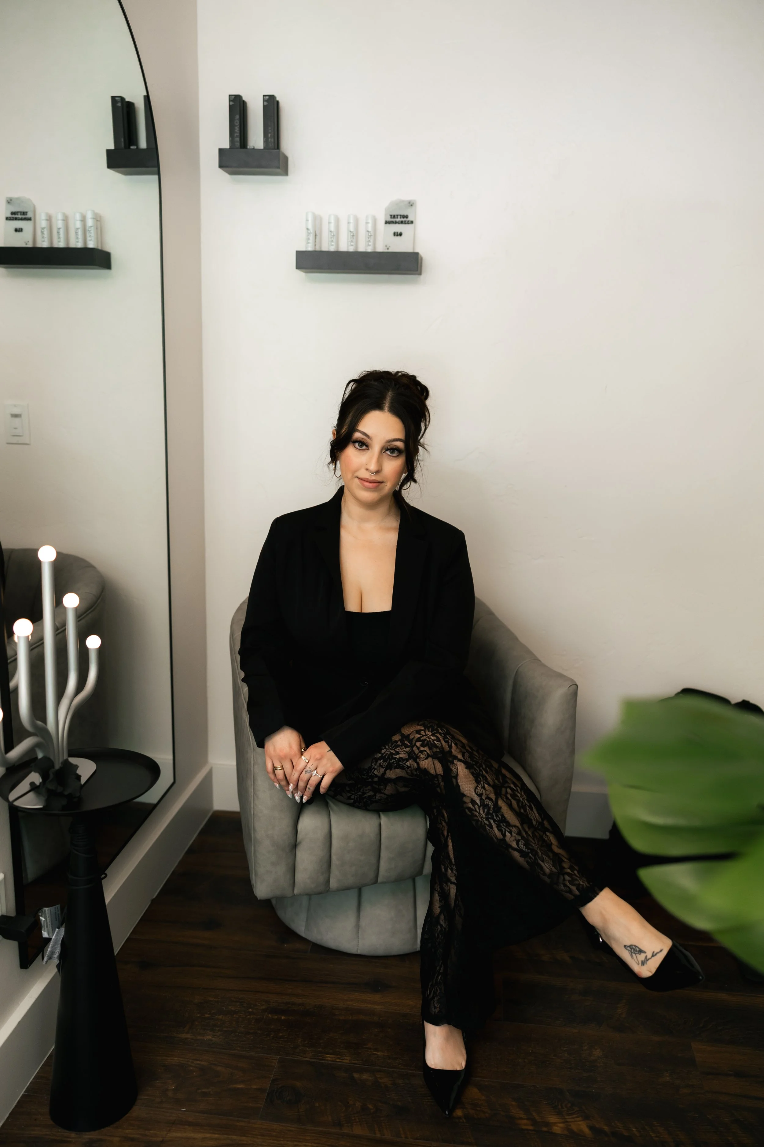 A woman with dark hair styled in an updo, wearing a black blazer and lace pants, seated on a gray armchair in a modern interior. She has tattoos on her foot and is wearing black heels. There are shelves with books and products on the wall behind her, and a large mirror reflecting part of the room. A black side table with candles and a menorah is nearby, and a plant is partially visible in the foreground.