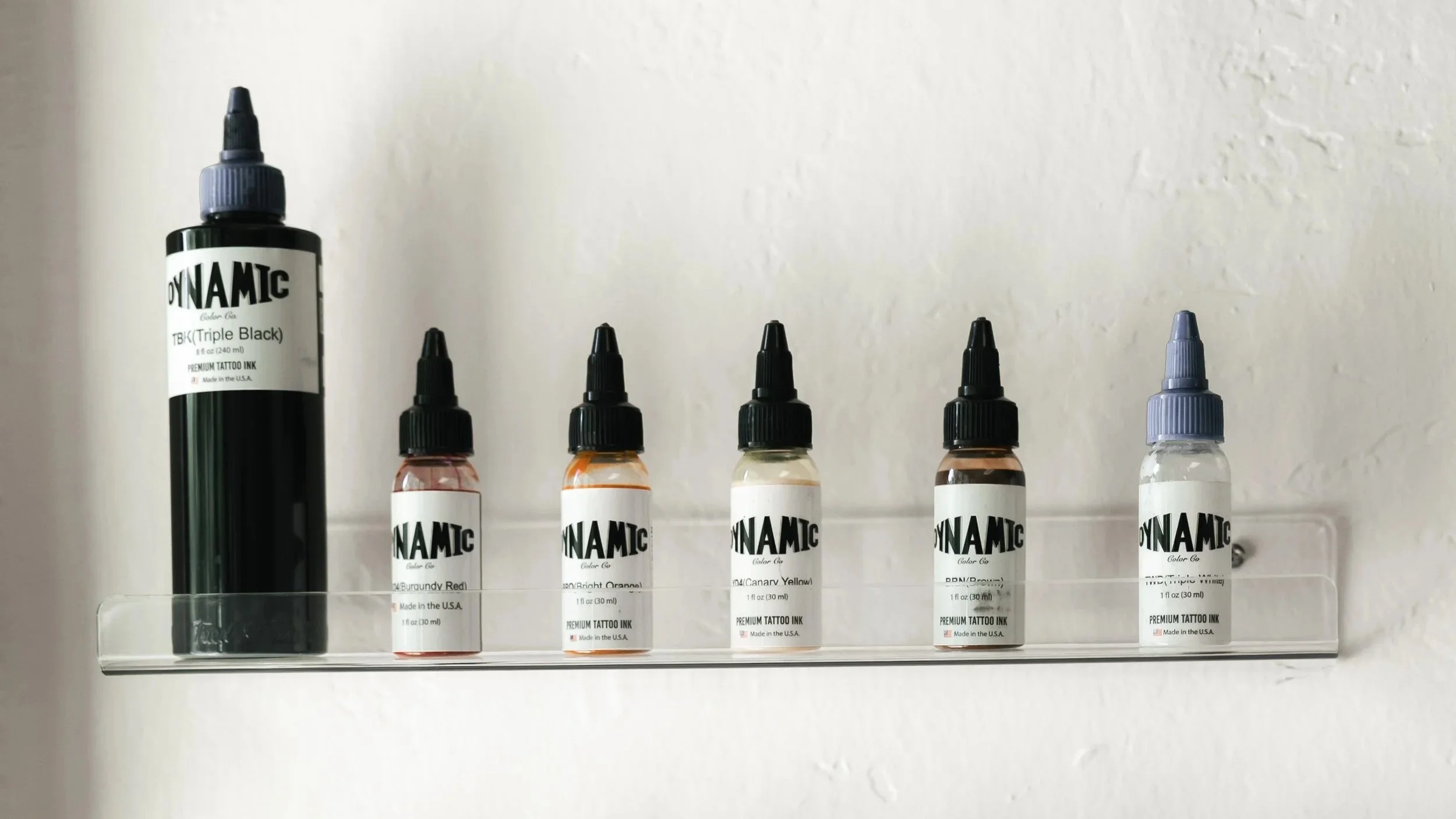 Display of tattoo ink bottles in various colors lined up on a clear shelf against a plain wall, with a large black bottle on the left and smaller bottles with black caps on the right.