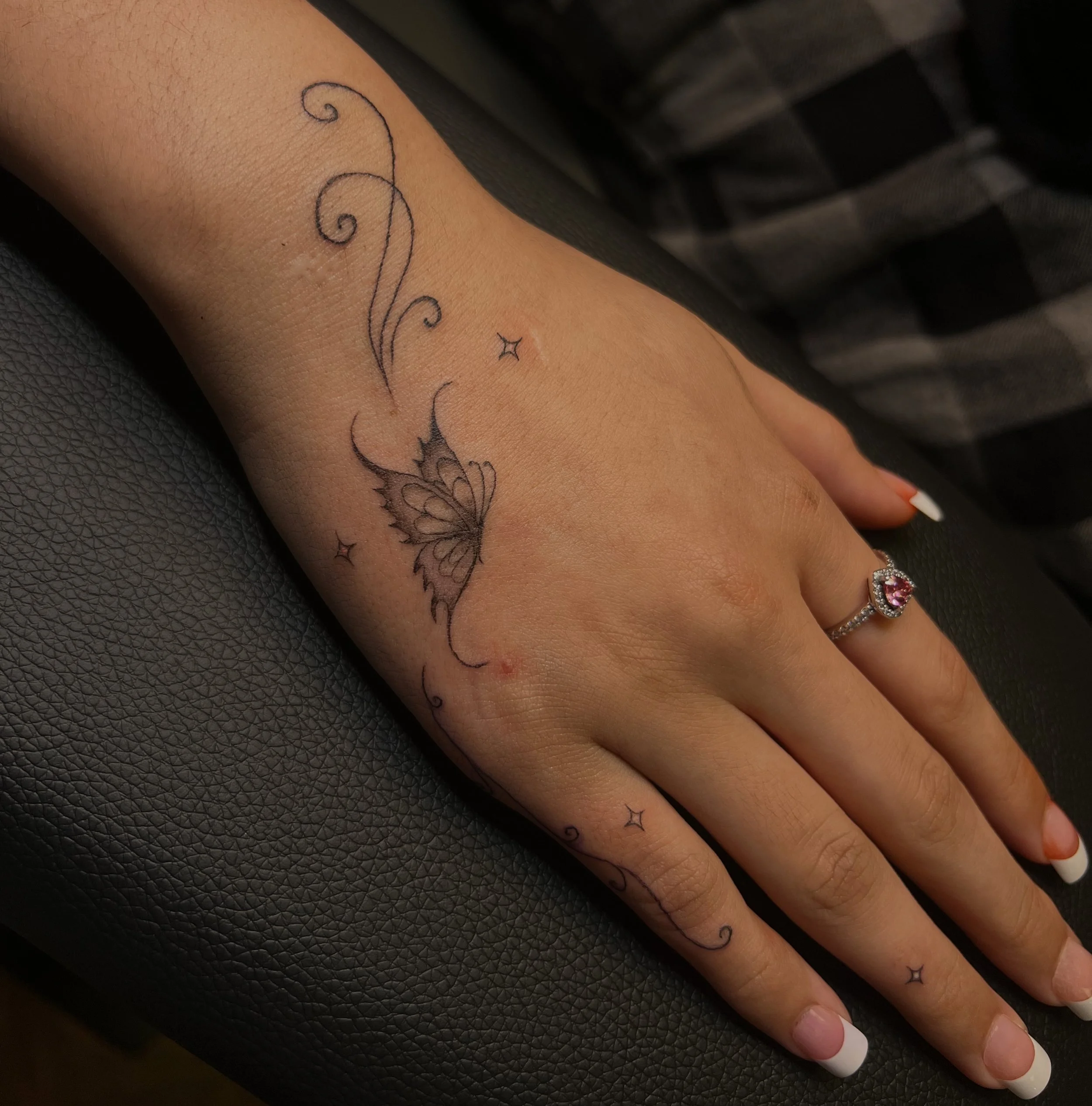 Close-up of a person's forearm showing a delicate black ink tattoo with swirling lines, stars, and a butterfly, while wearing a silver ring with a large pink gemstone on their finger.