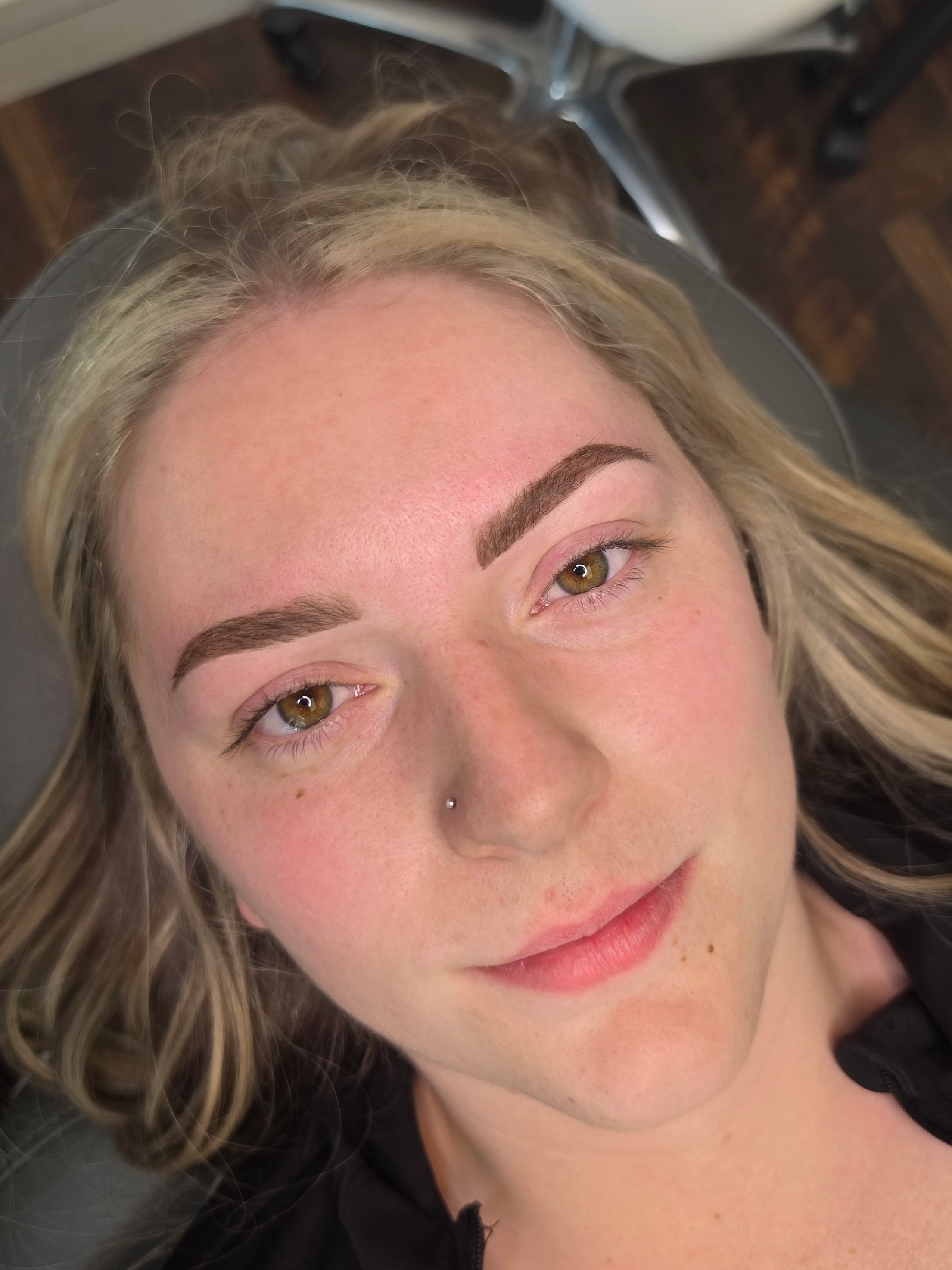A close-up of a woman's face, showing her green eyes, eyebrows, nose, and lips. She has blonde hair and is lying down, with part of a black jacket visible on her shoulder.