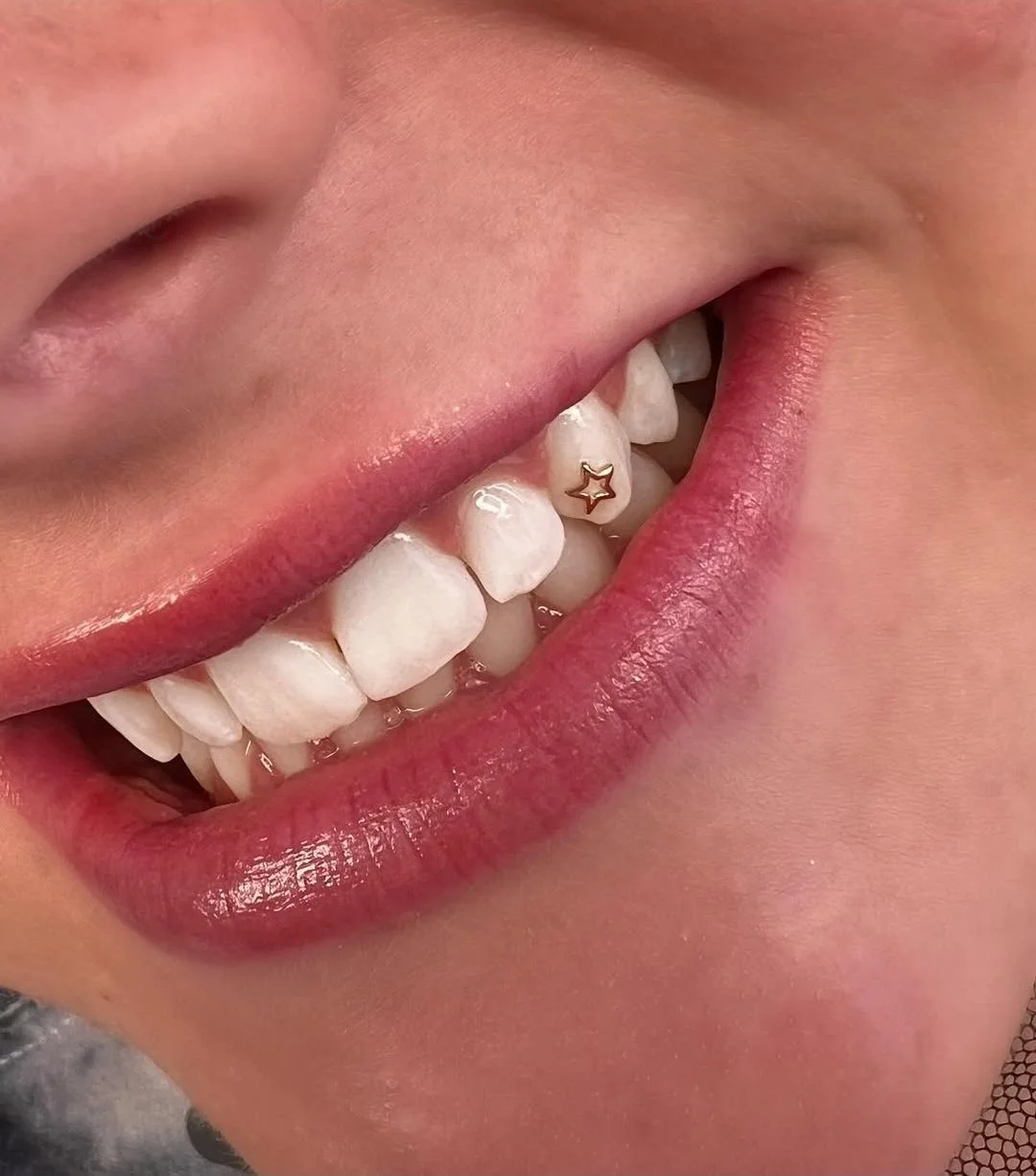 Close-up of a person's mouth with pink lipstick, showing white teeth with a gold star-shaped dental jewelry on one front tooth.