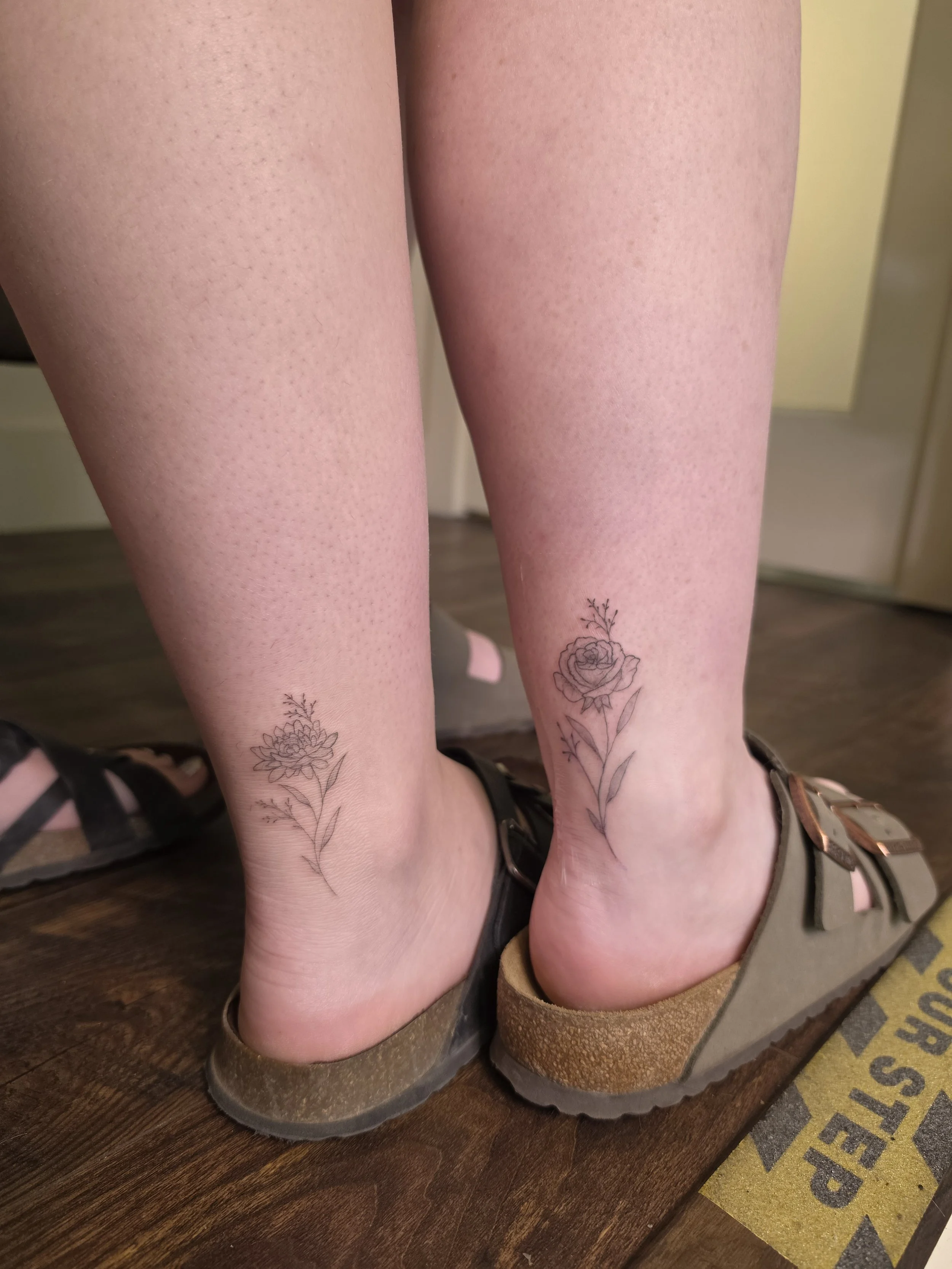 Close-up view of legs with tattoos of flowers, wearing shiny black and tan shoes, on a wooden surface with a yellow doormat nearby.
