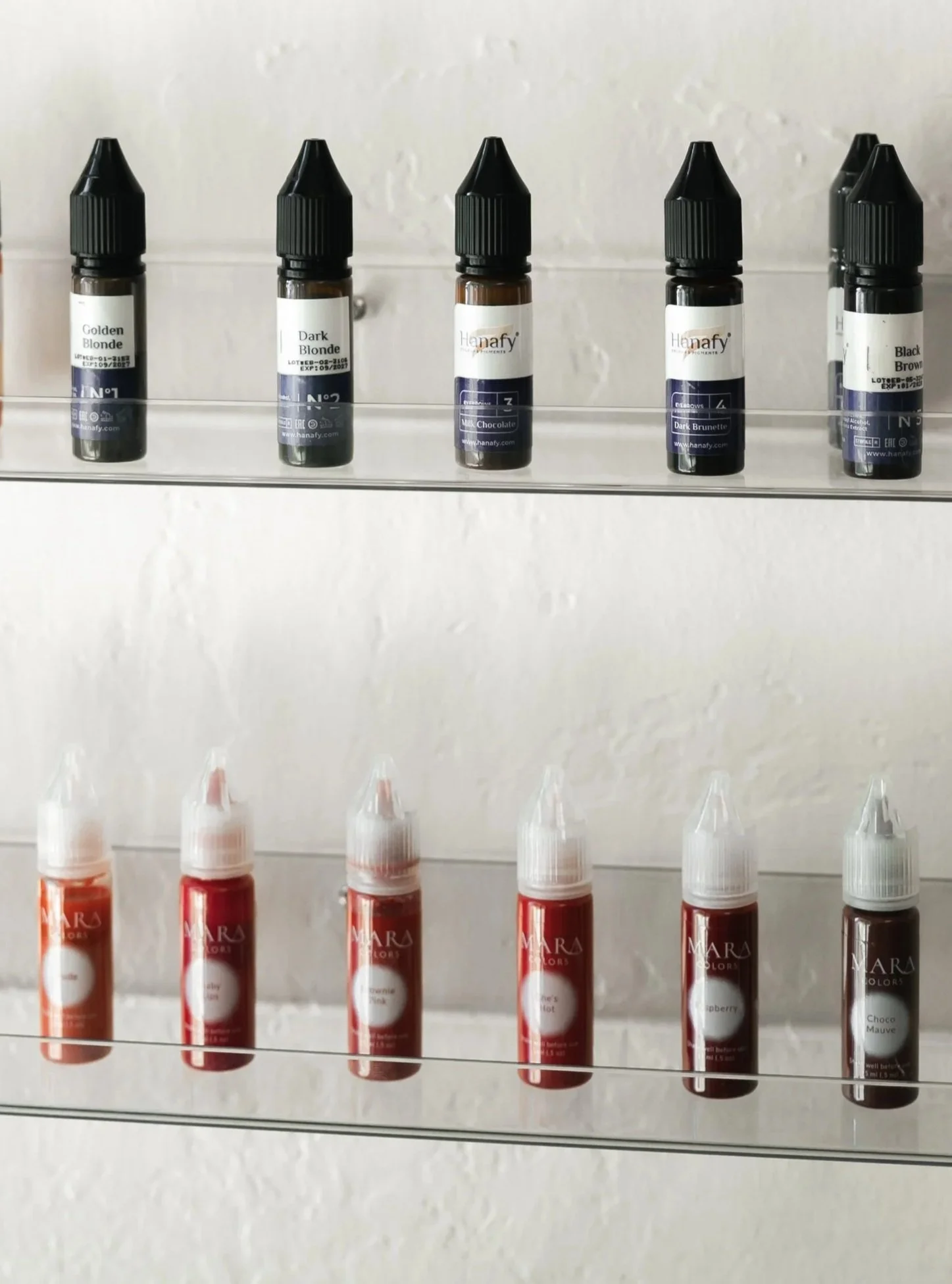 Small bottles of lip gloss or coloring products arranged on clear shelves, with some labels visible showing different shades like Golden Blonde, Dark Blonde, Milk Chocolate, Dark Brunette, and Black Brown.