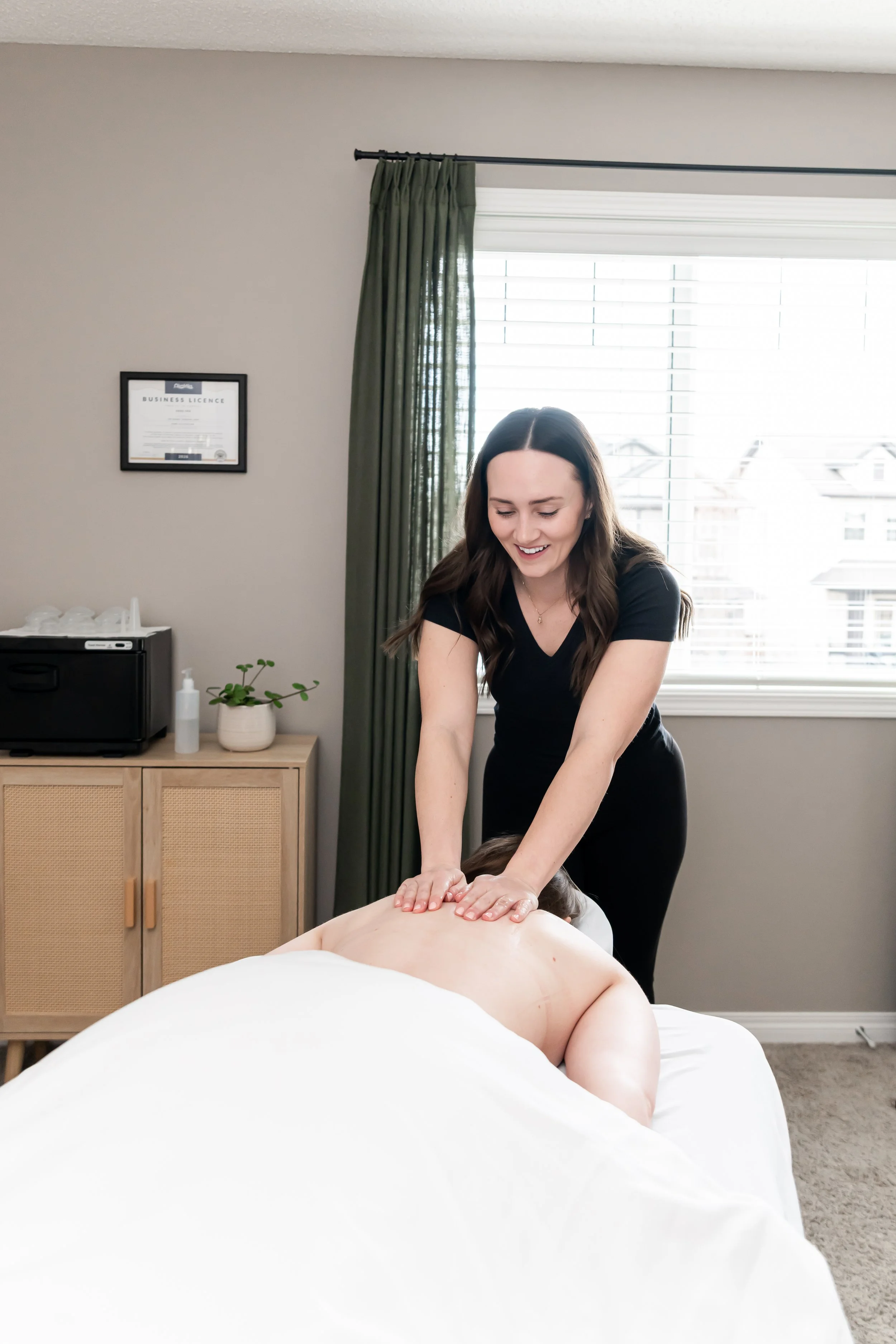 A woman giving a back massage to a person lying face down on a massage table in a well-lit room with a window, green curtain, and framed certificate on the wall.