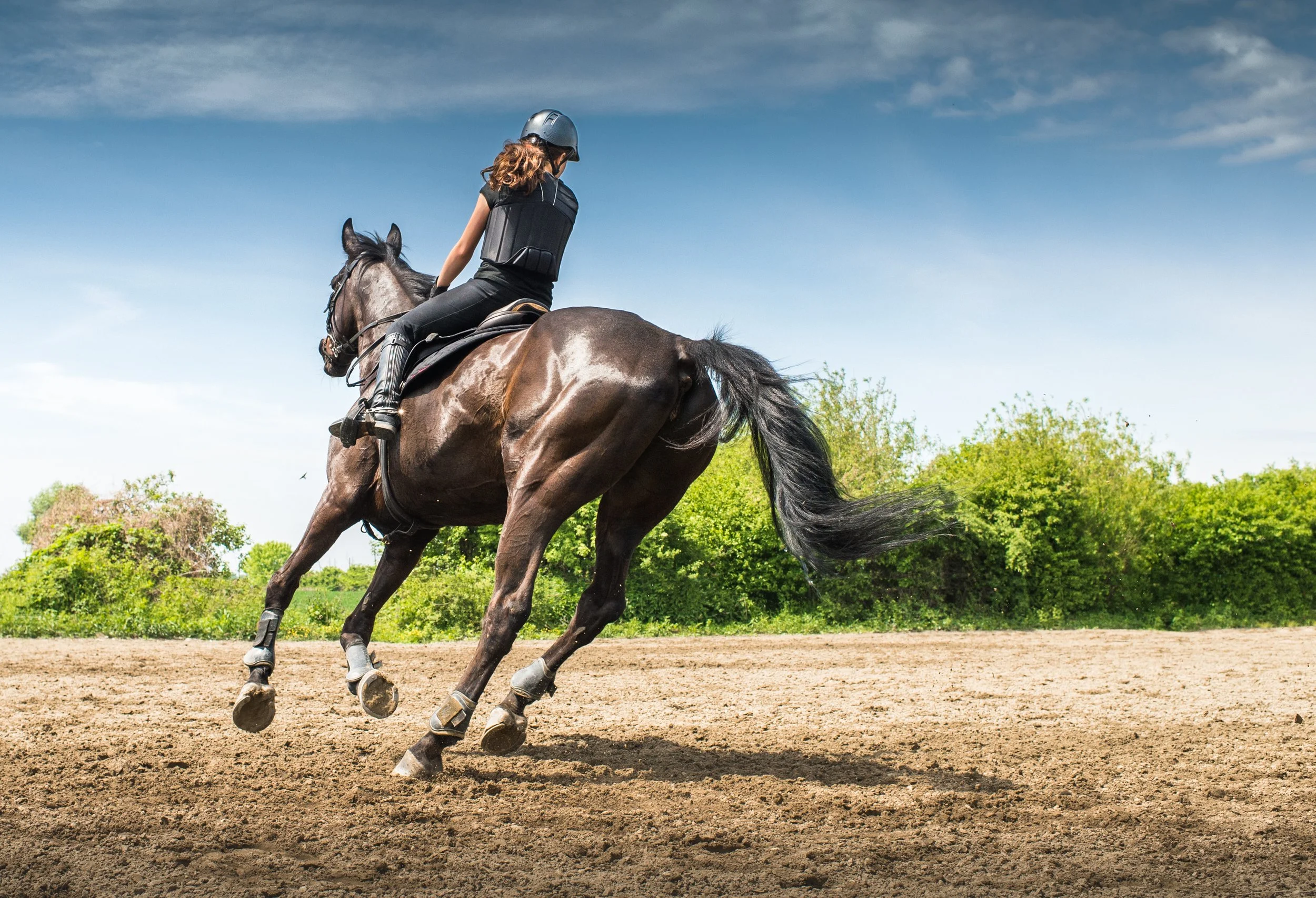 Une femme chevauche un cheval en pleine course sur une piste en terre, sous un ciel bleu avec quelques nuages et des arbres verts en arrière-plan.