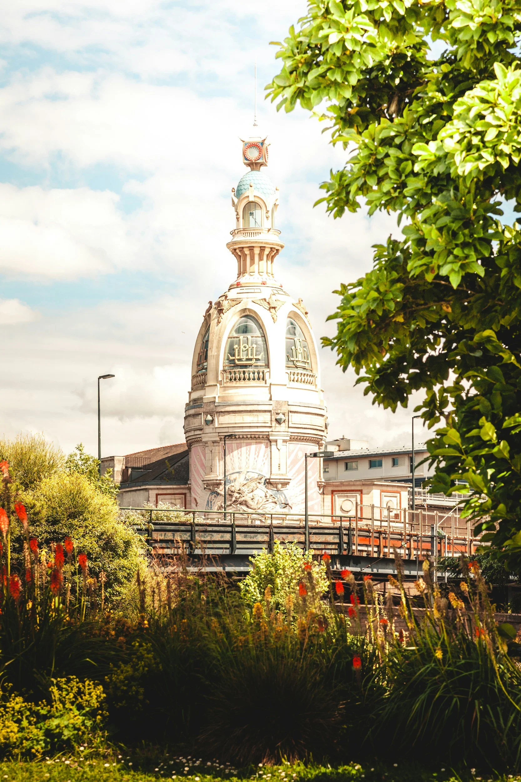 Une image représentant une tour ornée de détails architecturaux, partiellement encadrée par des arbres et des plantes dans un jardin, sous un ciel partiellement nuageux.
