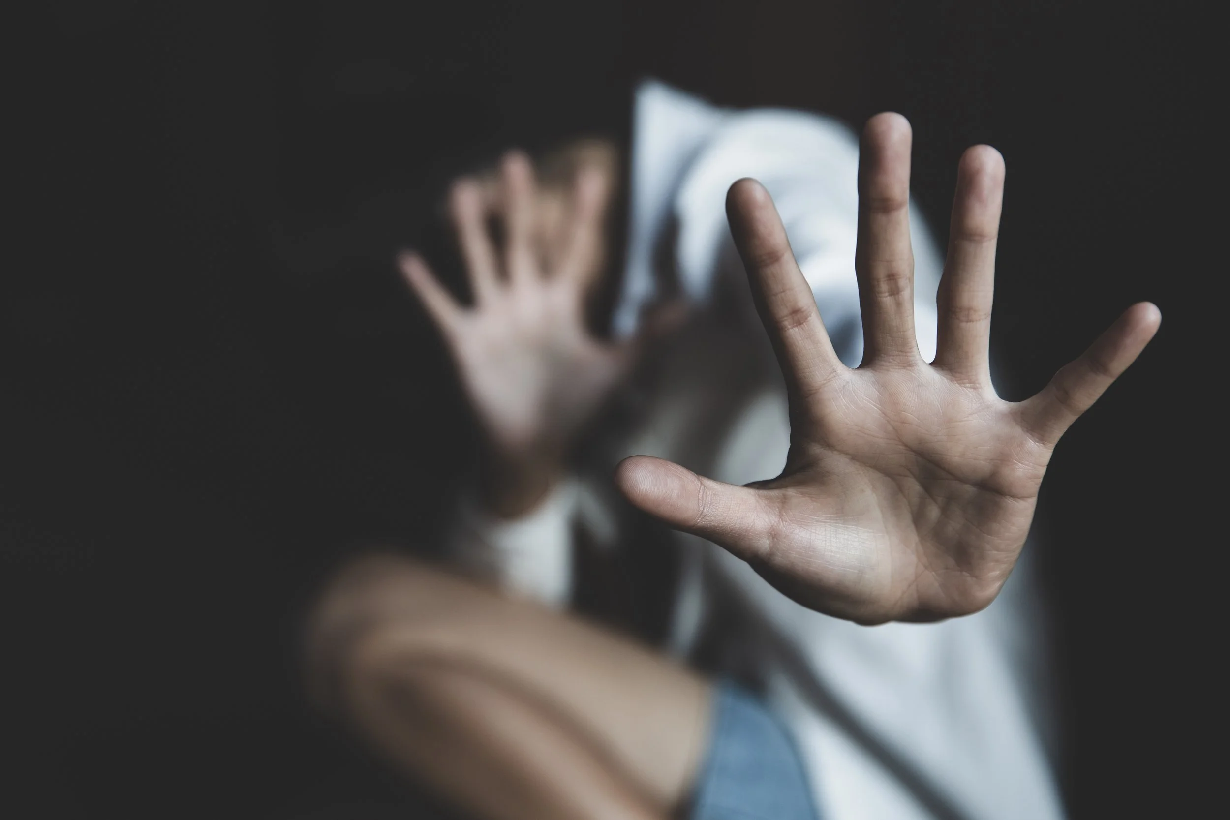 Main human hand levée avec doigts écartés en premier plan, personne floue derrière avec main sur visage, en milieu sombre.