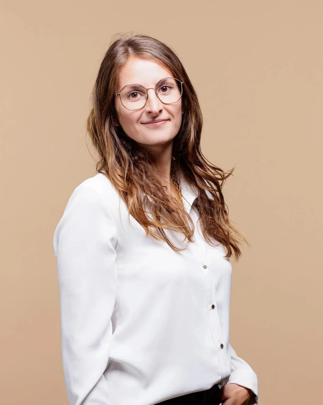 Une femme aux cheveux bruns, portant des lunettes et une chemise blanche, pose face à un fond beige.