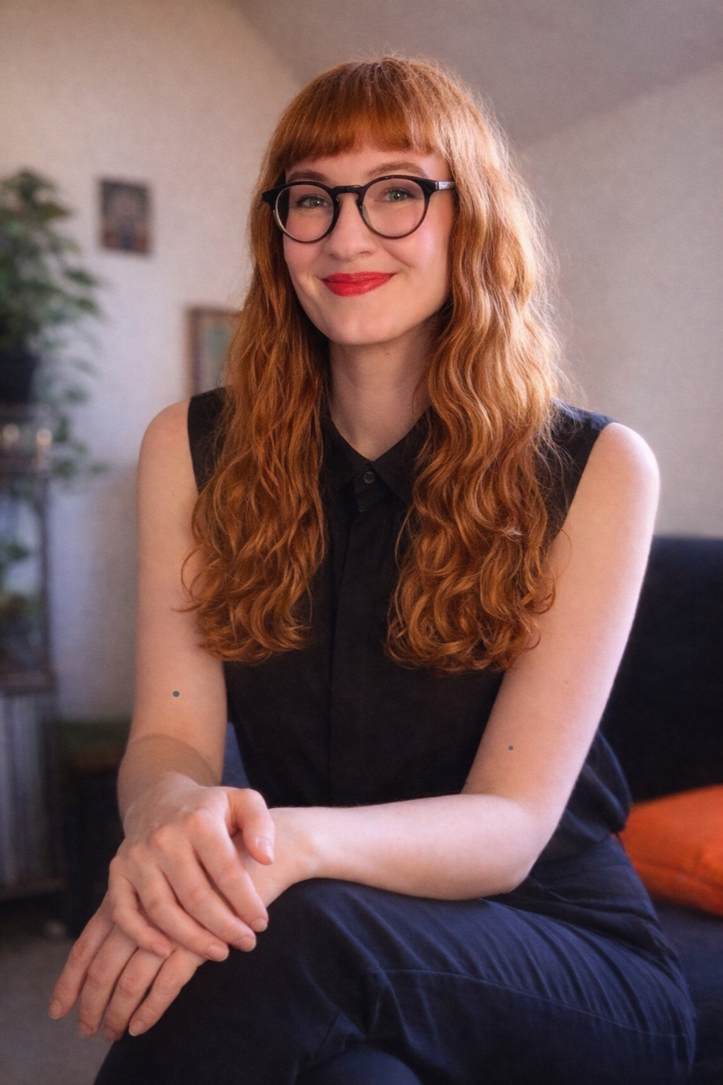 A confident young woman with long, curly red hair, wearing glasses and a sleeveless black top, sitting indoors with a warm smile.