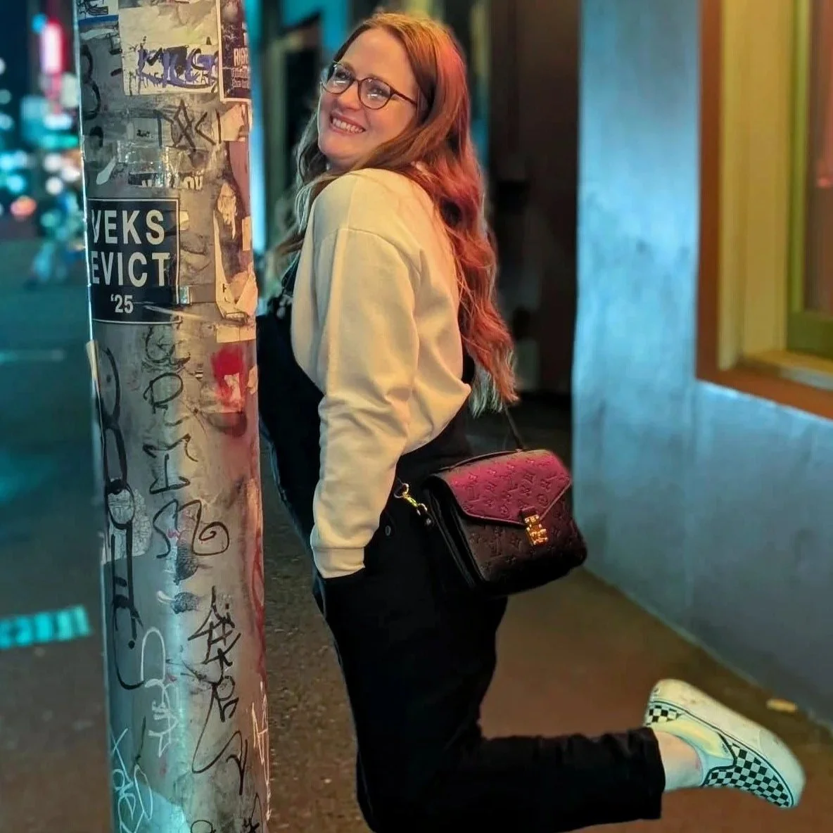 A young woman with long red hair, glasses, and a beige jacket leaning against a graffiti-covered pole on a city street at night. She is smiling, carrying a pink and black handbag, and wearing checkerboard slip-on shoes.
