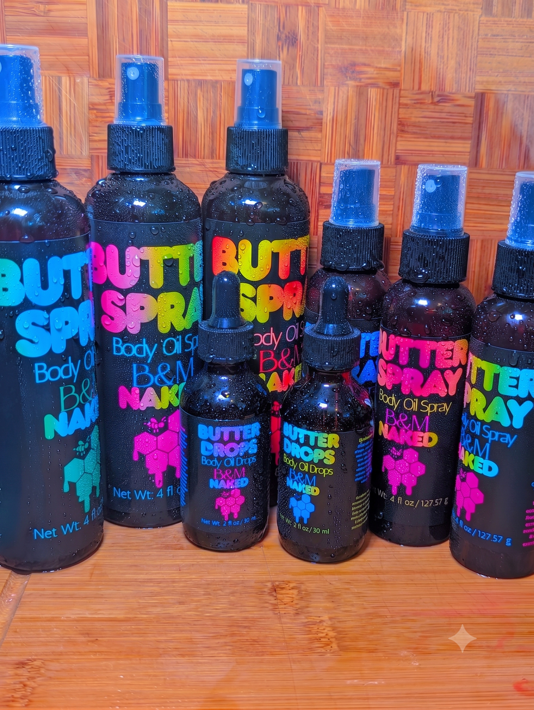 Set of black bottles labeled "BUTTER SPRAY" with colorful text and graphics, arranged on a wooden surface against a wooden background, condensation visible on the bottles.