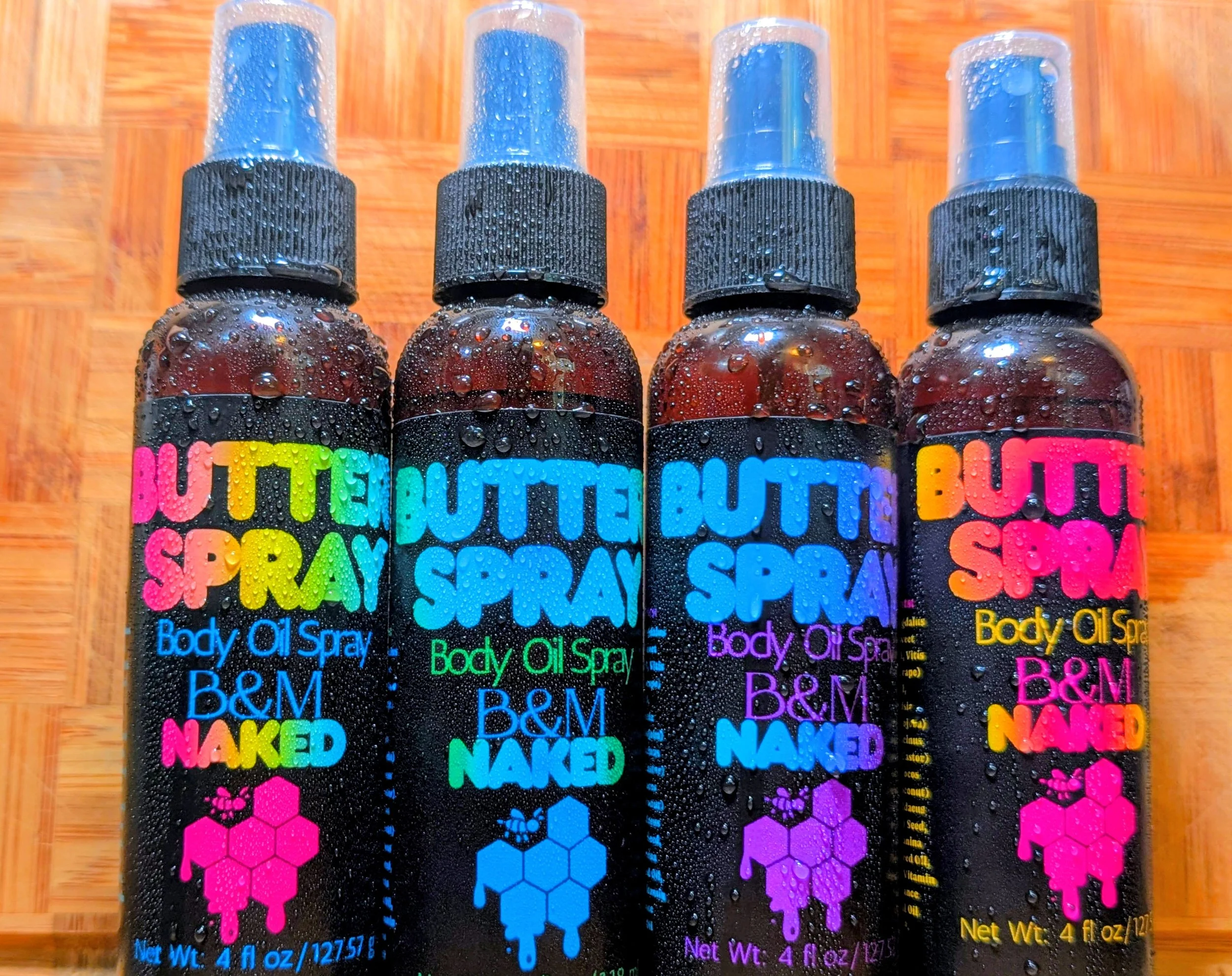 Four bottles of body oil spray with colorful labels and condensation droplets on the surface, placed on a wooden surface.