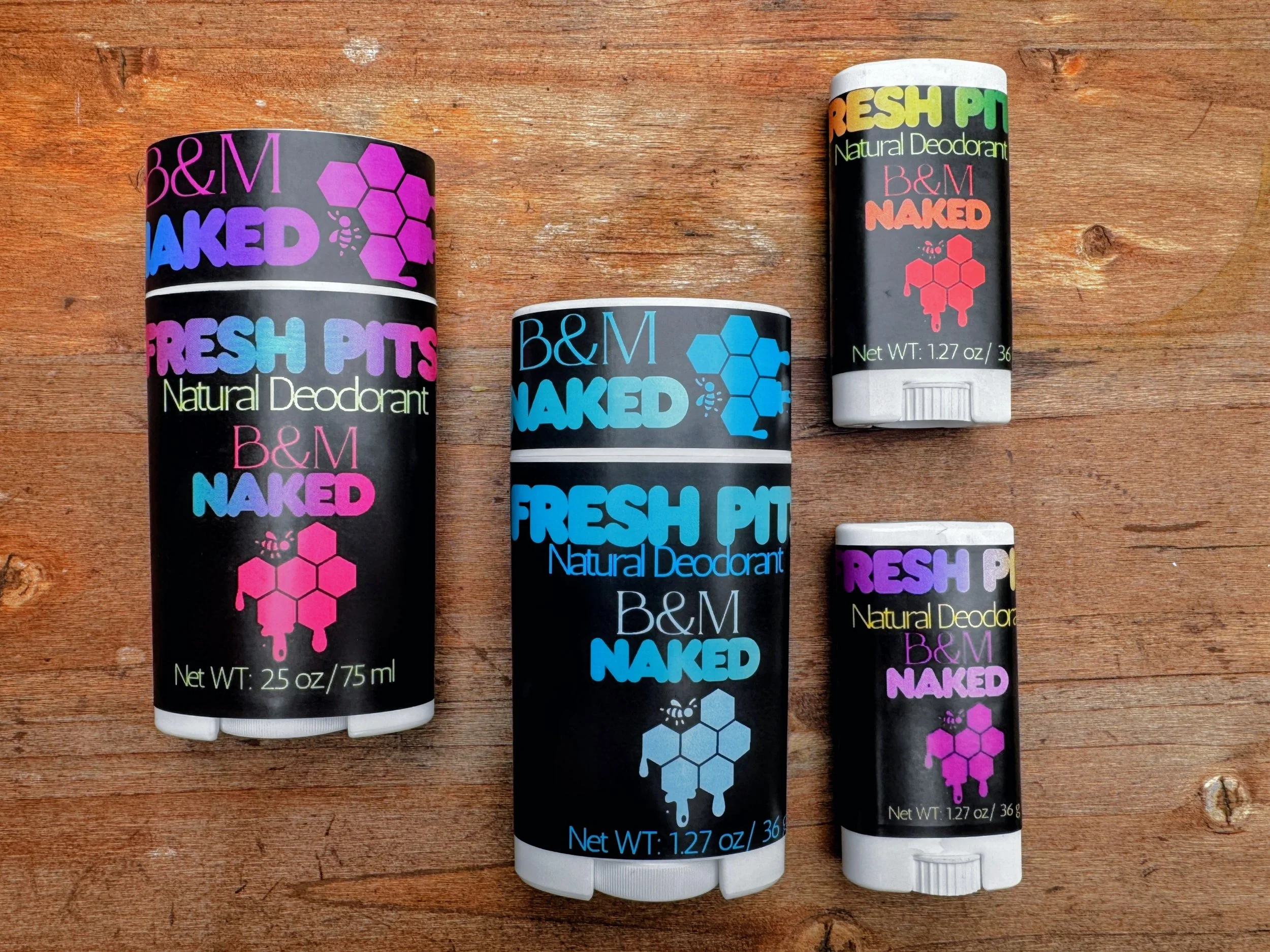 Three sticks of B&M Naked natural deodorant on a wooden surface with visible wood grain and knots. The deodorants are black with colorful packaging and text.