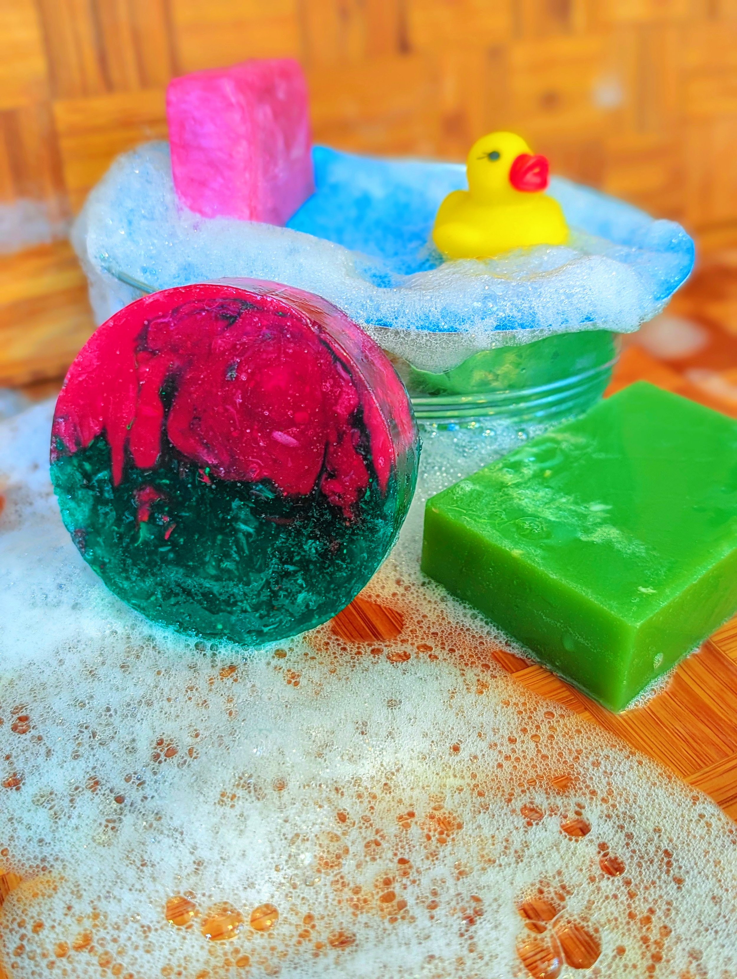 Colorful bath bombs in various shapes including a yellow rubber duck, a green rectangle, pink, blue, and red round shapes, set in soapy water with bubbles and foam.