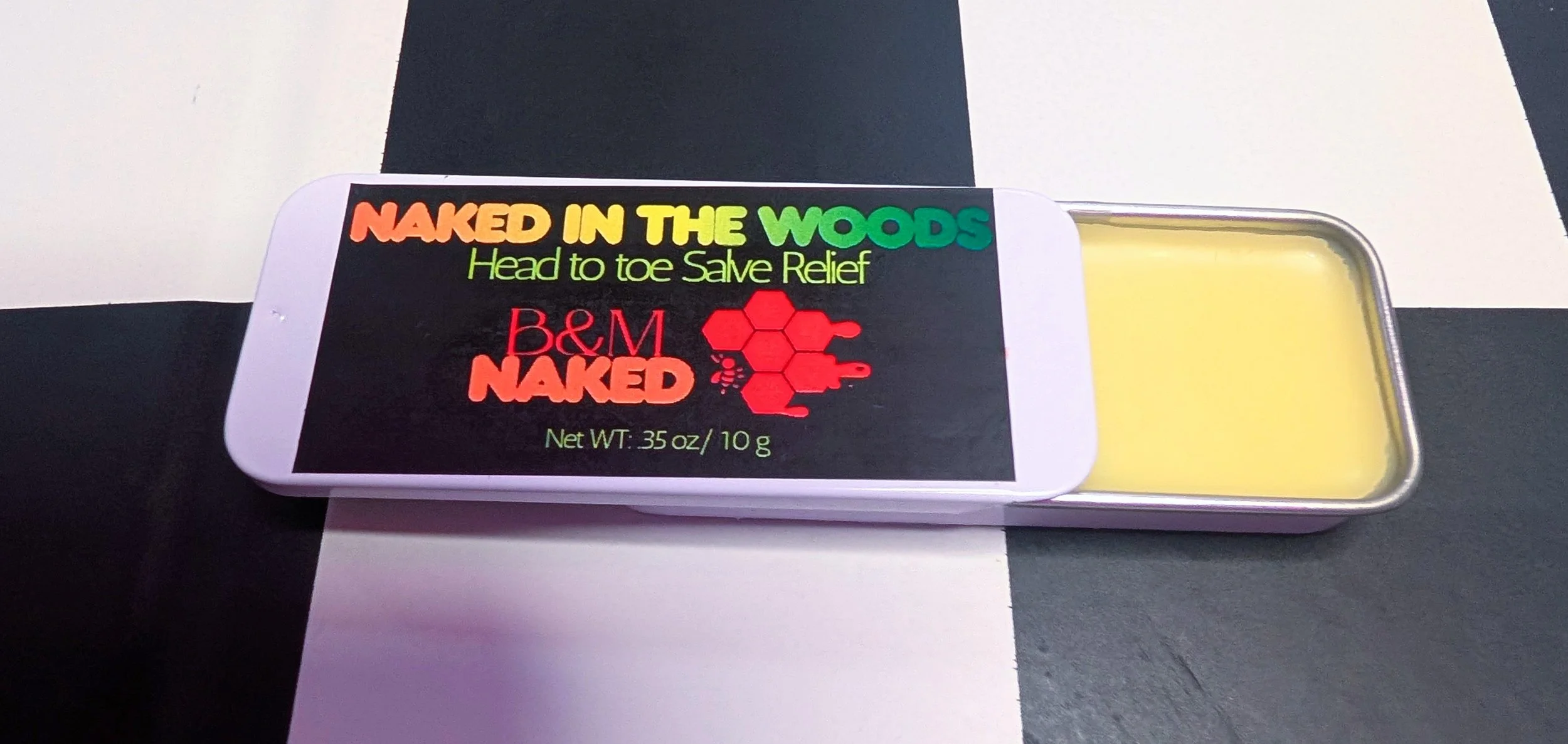 A bar of salve labeled "NAKED IN THE WOODS," with green, yellow, orange, and red text, and a red hexagon logo. The salve is in a metal tin with a yellowish balm inside, placed on a black and white checkered surface.