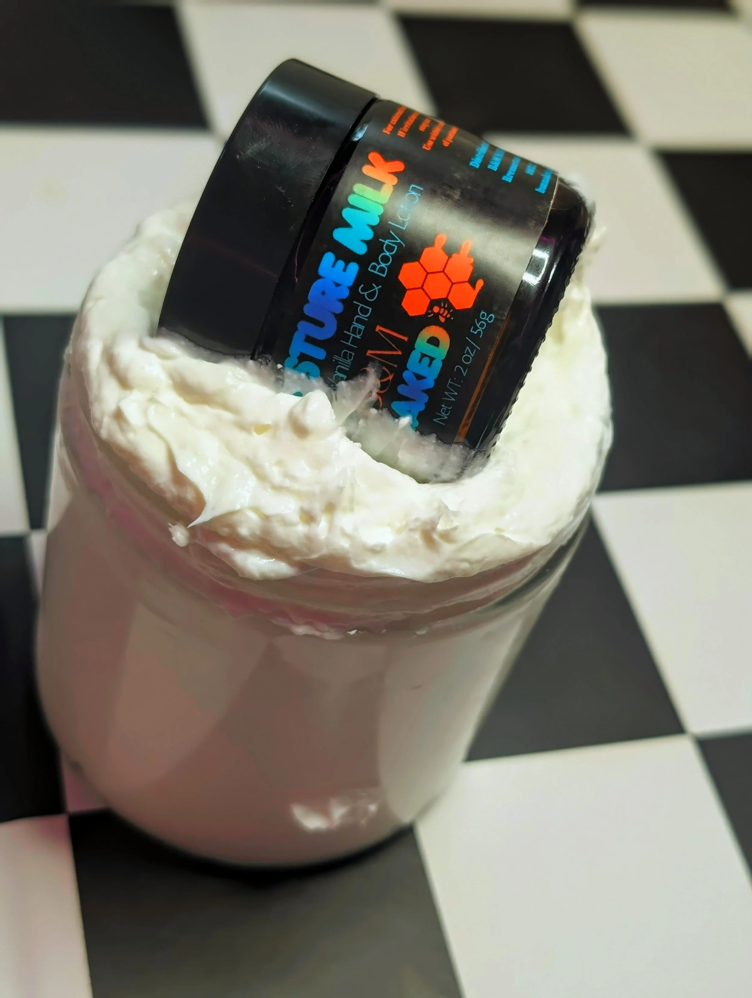 A small black container labeled 'Strawberry and Body Lotion' with a honeybee illustration, placed inside a glass filled with whipped cream, on a black and white checkered surface.