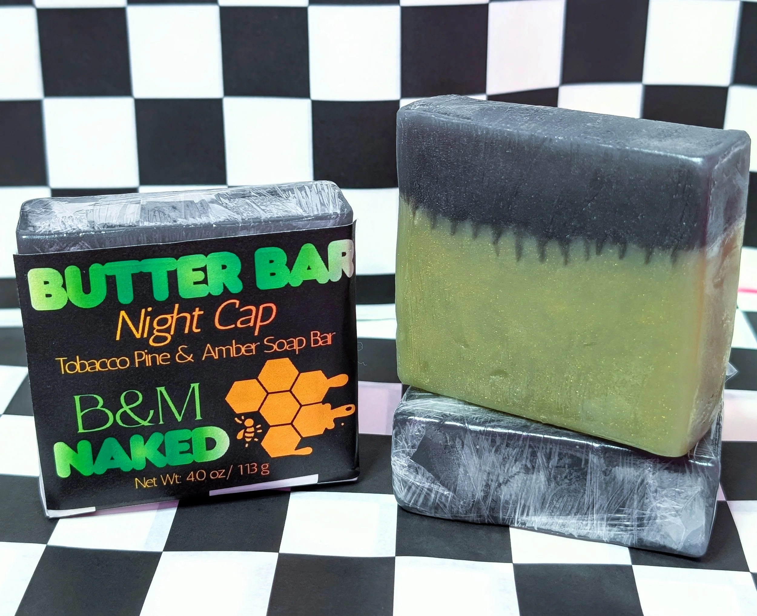A package of Butter Bar Night Cap soap with a black background and green text, next to a large rectangular bar of soap that is gray and yellow, with a textured green base, all on a black and white checkered surface.