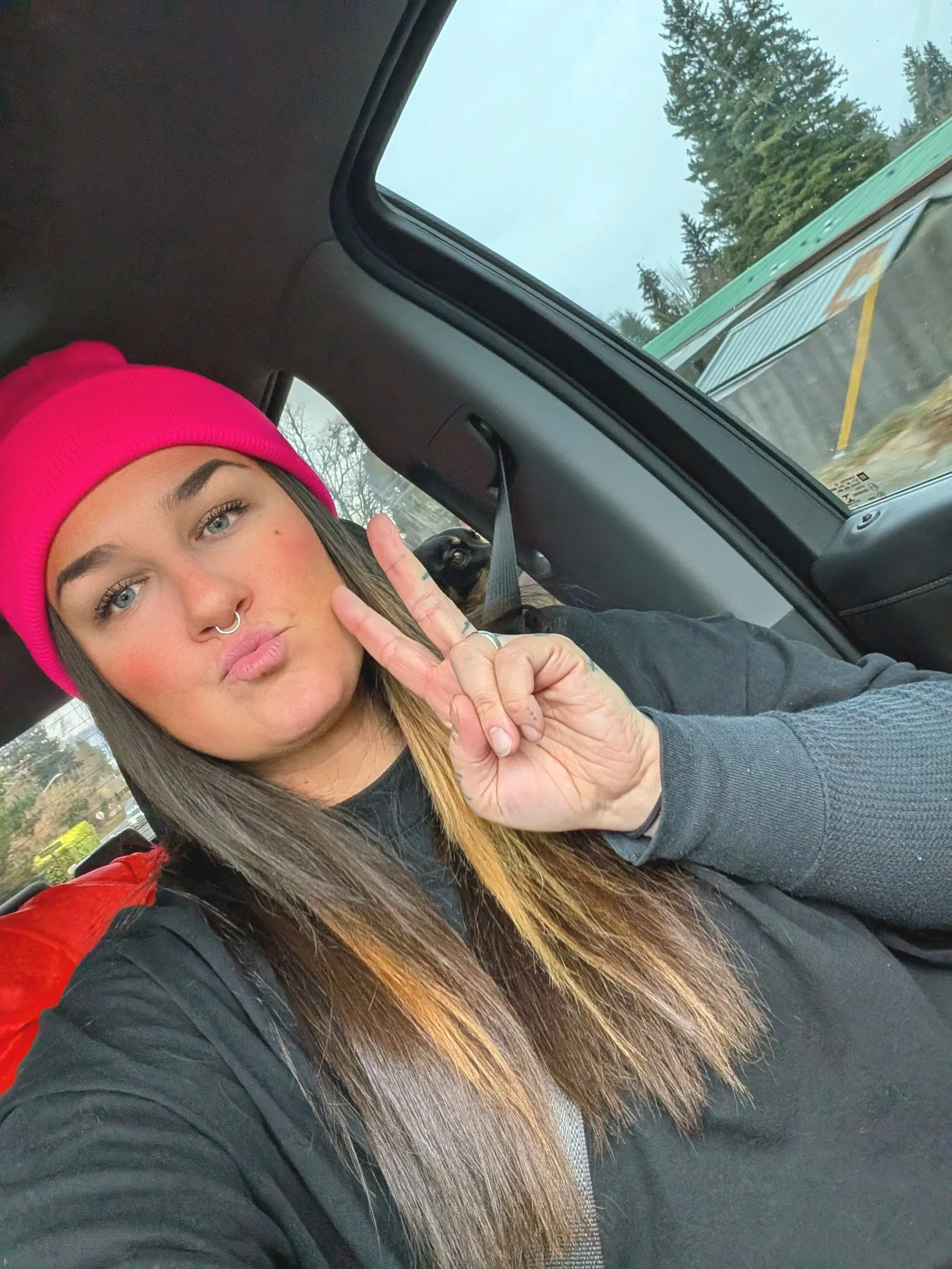 A young woman with long, straight brown hair and light-colored eyes, wearing a bright pink beanie and black jacket, sitting in a car, making a peace sign with her right hand. A small black dog is visible in the background, with trees and a building seen through the car window.