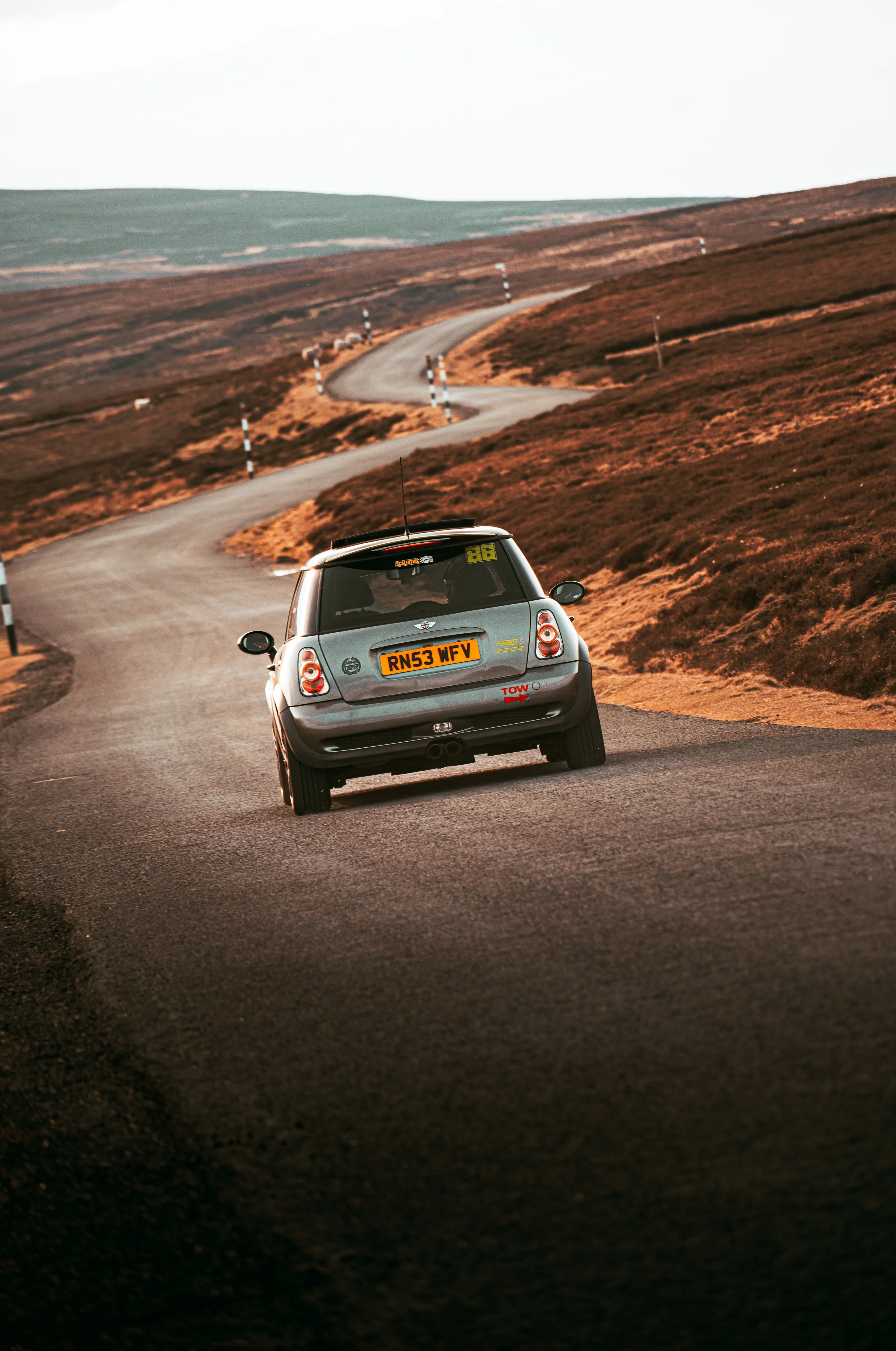 A silver car driving on a winding road through a hilly landscape with brown and green vegetation.