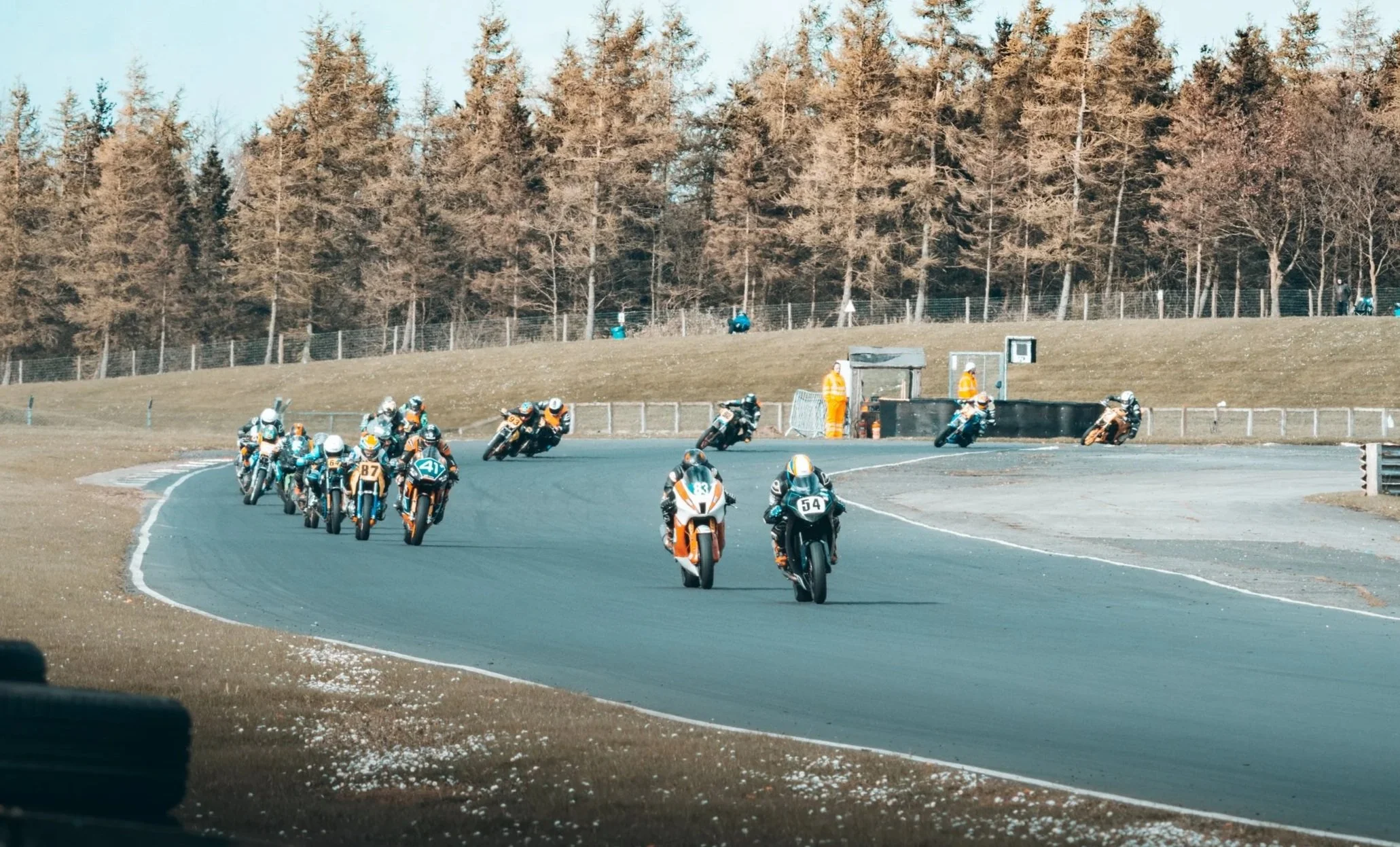 Motorcycle race on a curved track surrounded by trees with a fence and some officials watching.
