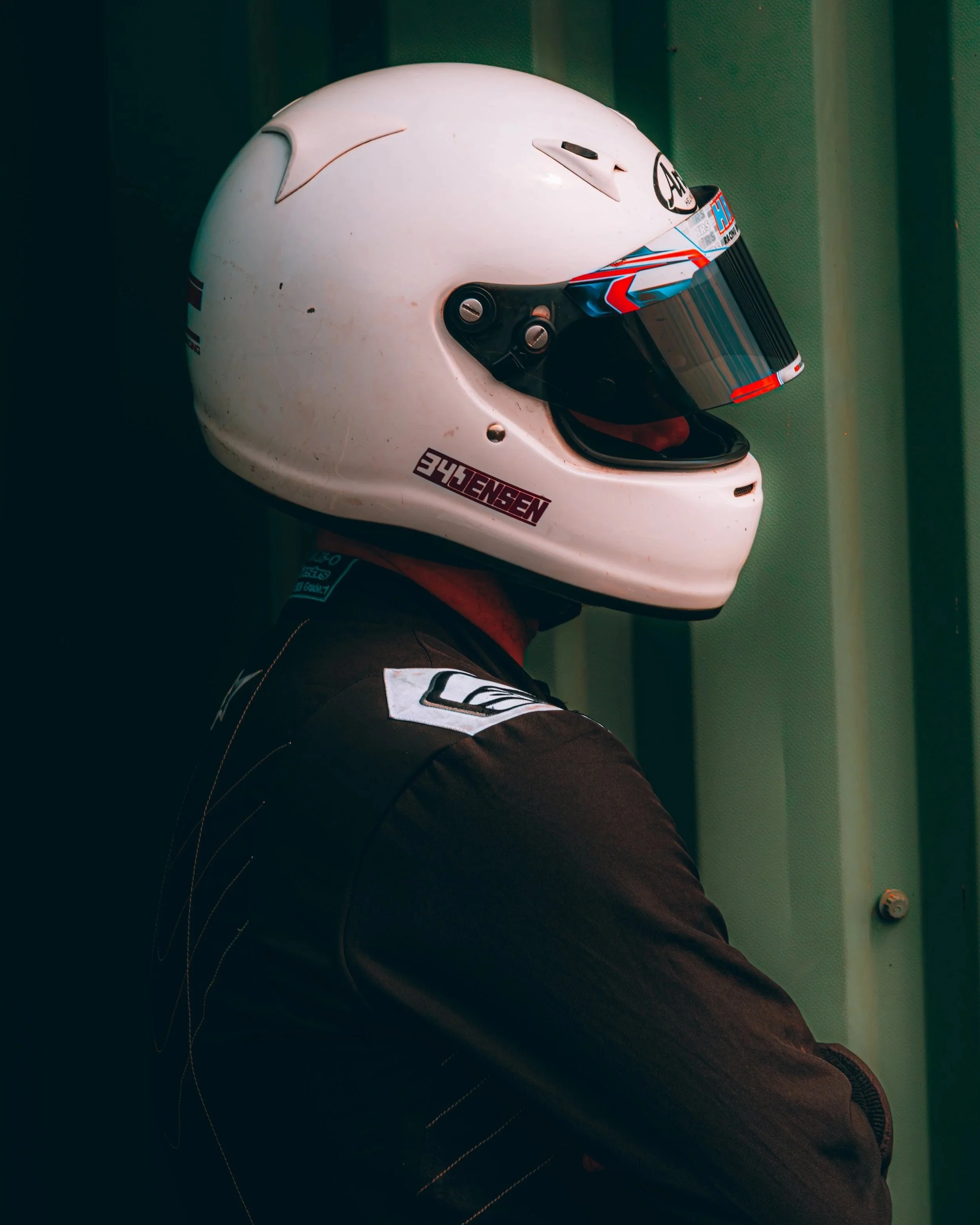 A white racing helmet with a colorful visor and the brand name 'HJC' on the front. The helmet is placed on the person's lap in a racing suit, with a green textured wall in the background.