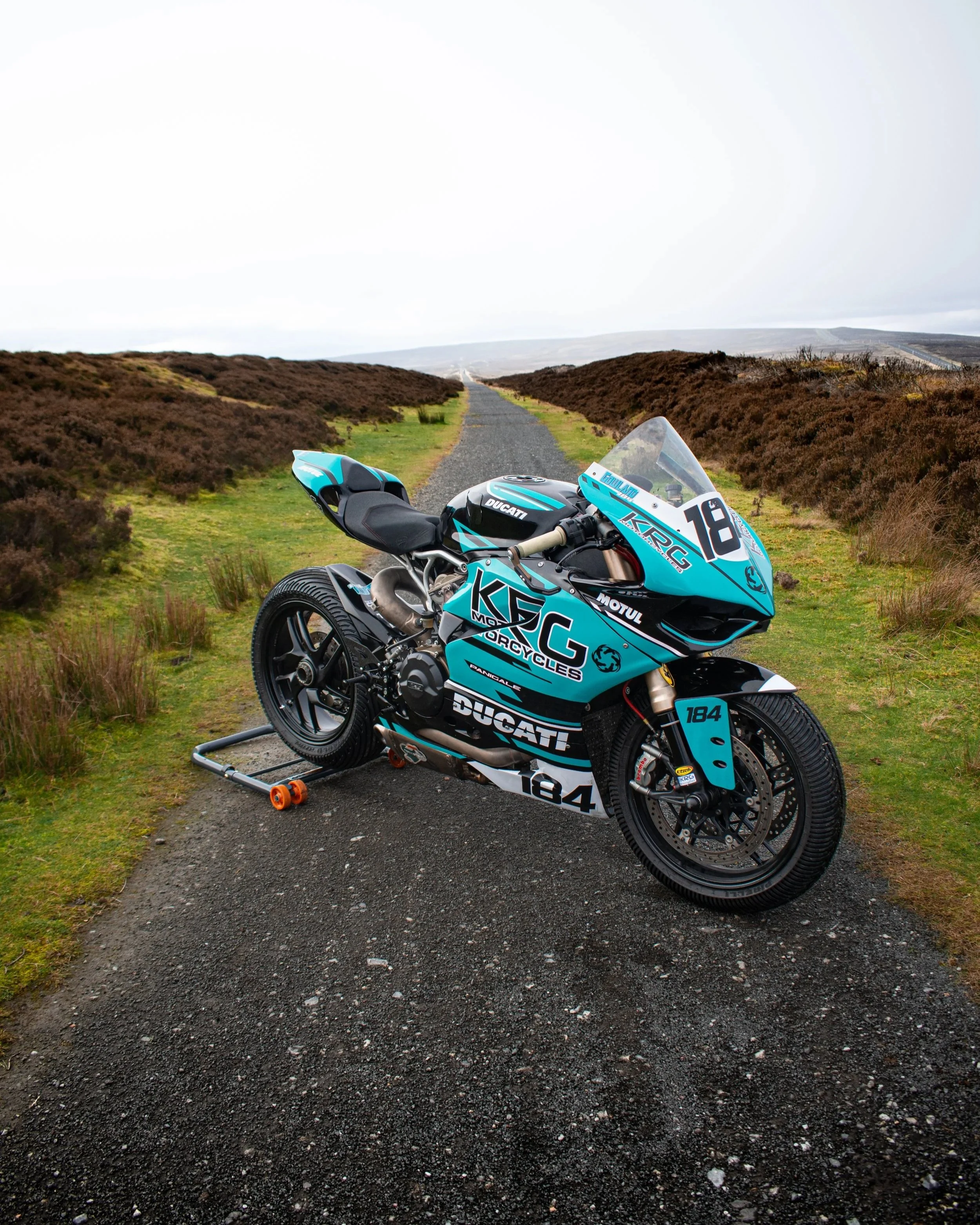 A blue and black Ducati motorcycle with racing decals is parked on a gravel path in a rural landscape with brown bushes and hills in the background.