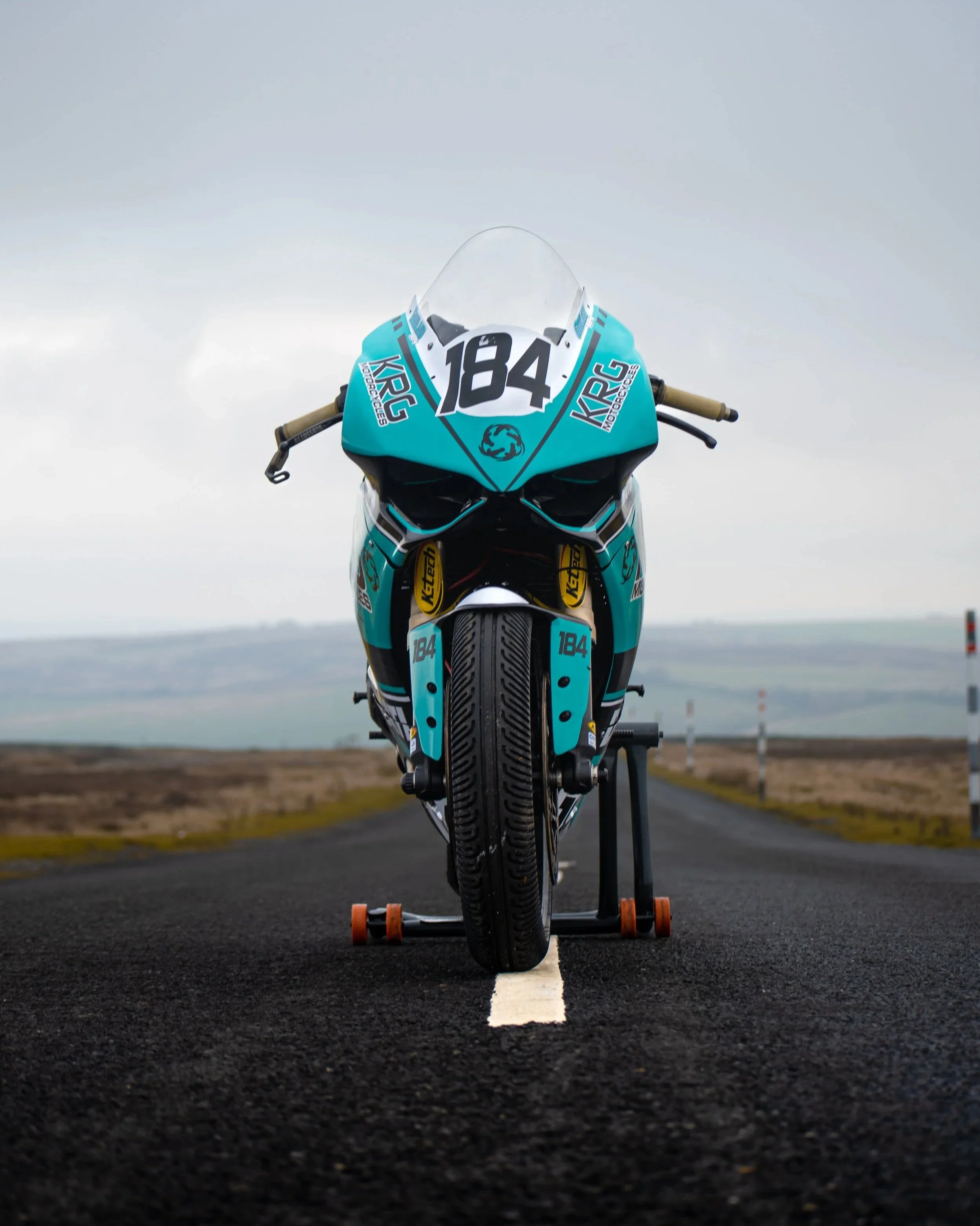 A racing motorcycle with the number 184 on it, positioned on an asphalt track with a white line, supported on a stand, under an overcast sky.