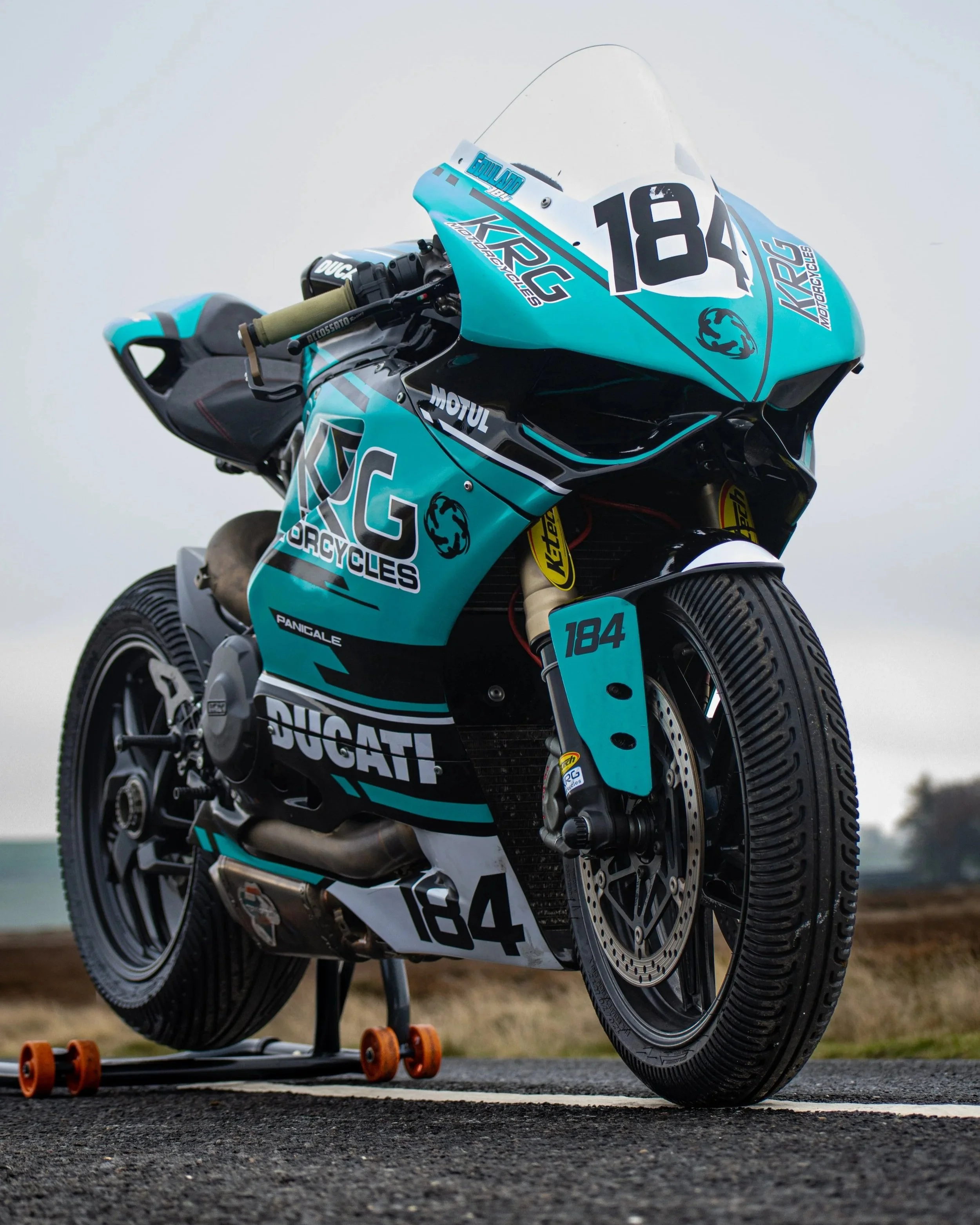 A racing motorcycle with dark tires, teal and black body, and a number 184. It is positioned outside on a textured gray surface during daytime, with a blurred background. The motorcycle has branding from Ducati and Kawasaki, and is mounted on a small