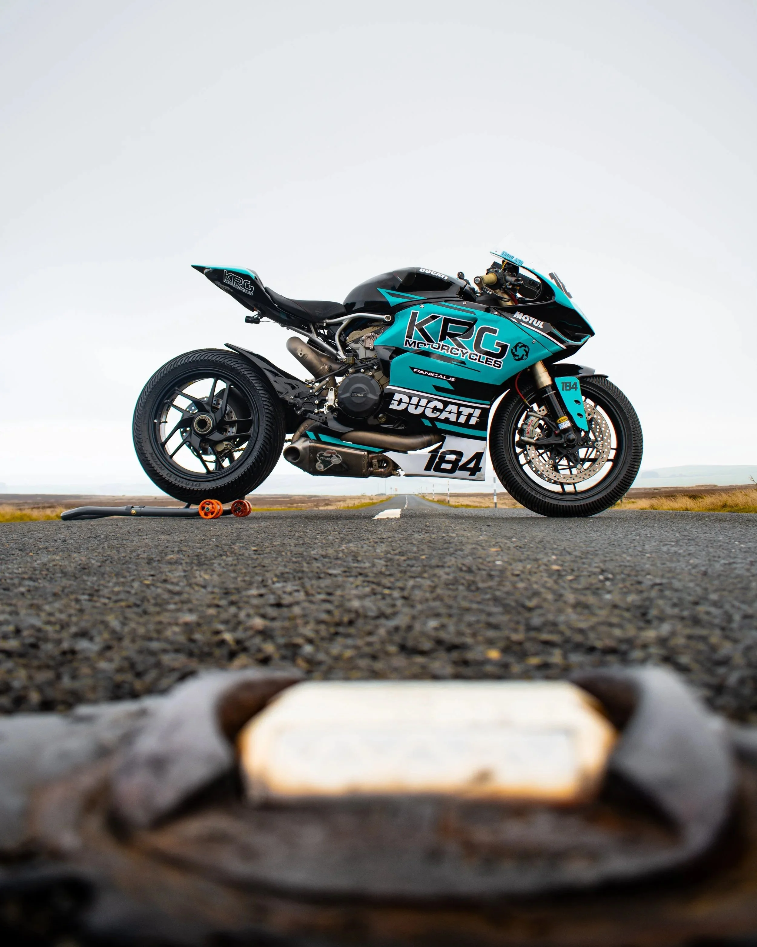 Close-up view of a racing motorcycle on a road, with the focus on the tire and wheel at the foreground and the motorcycle's bodywork featuring branding and racing number '184' in the background.