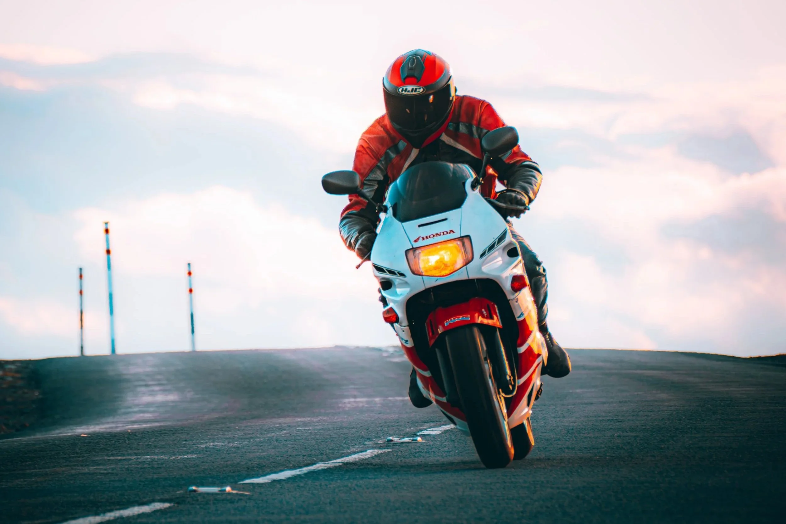 A motorcyclist wearing a red helmet and jacket performing a wheelie on a white and red Honda motorcycle on a racetrack during sunset.