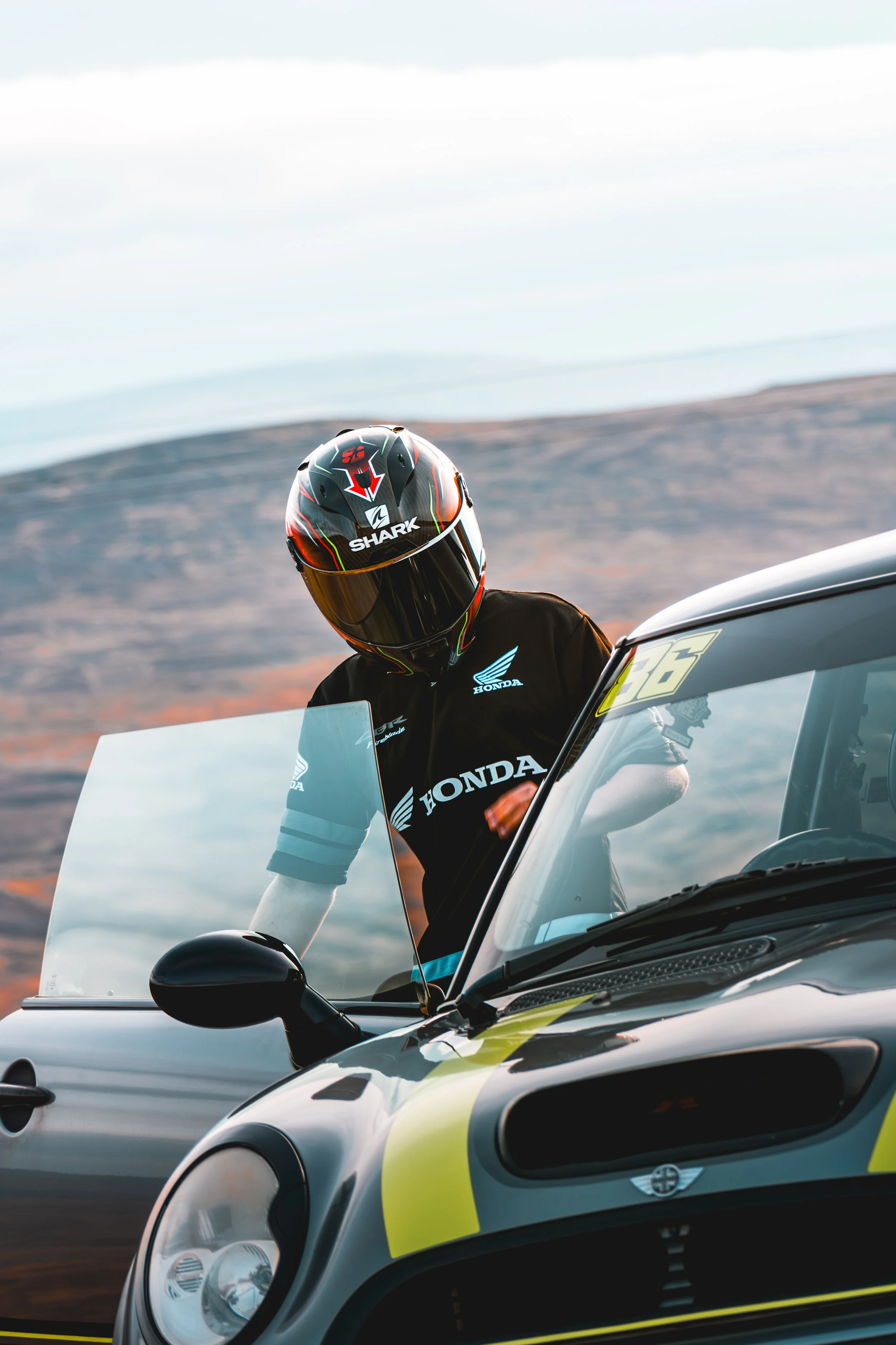 A person wearing a black helmet with red and white graphics, and a black jacket with Honda branding, is standing outside next to a vehicle with a mountain and cloudy sky in the background.