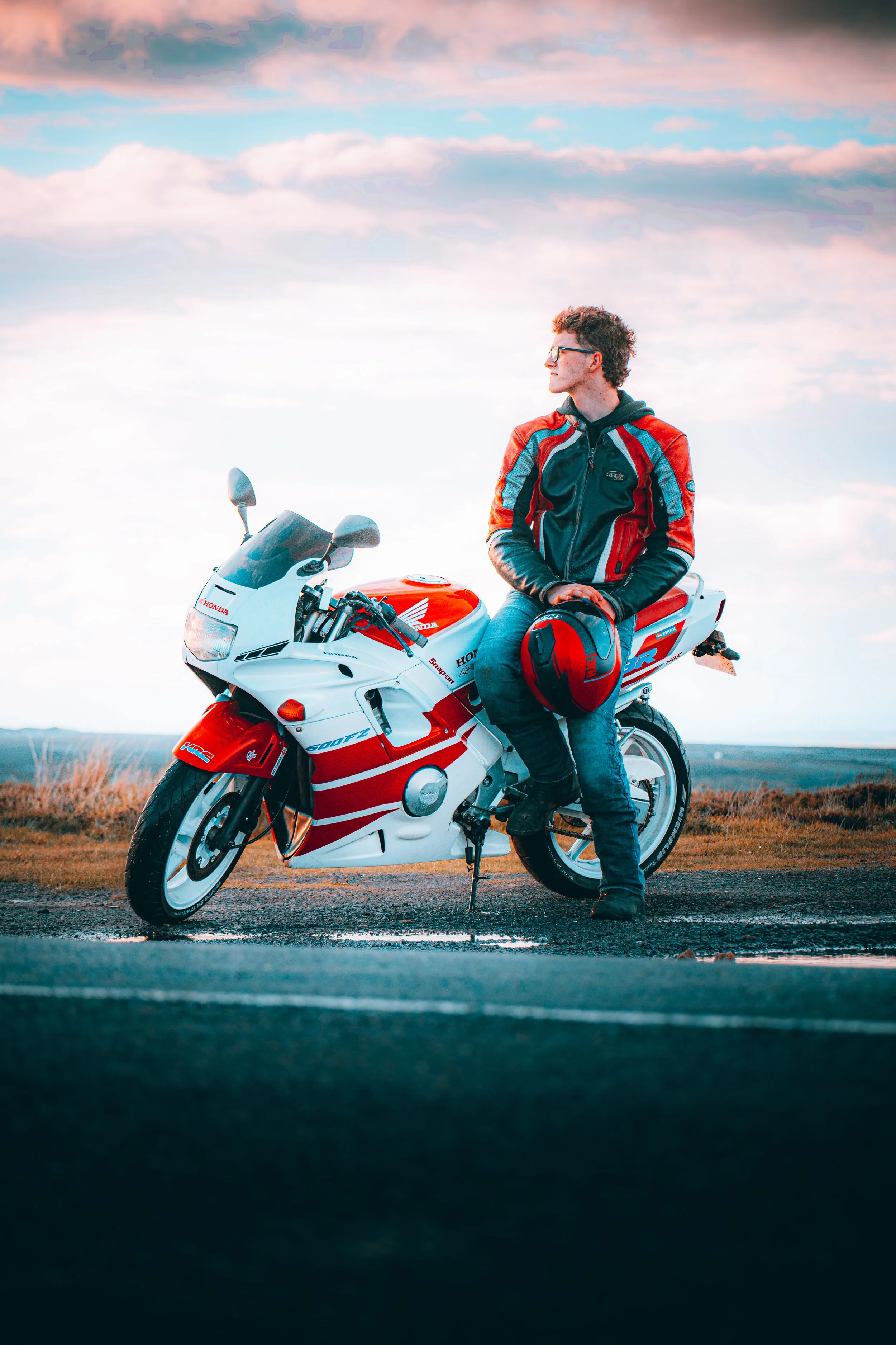 A man wearing a motorcycle jacket and jeans sitting on a white and red Honda motorcycle on a road, holding a red helmet, with an open sky and clouds in the background during sunset.