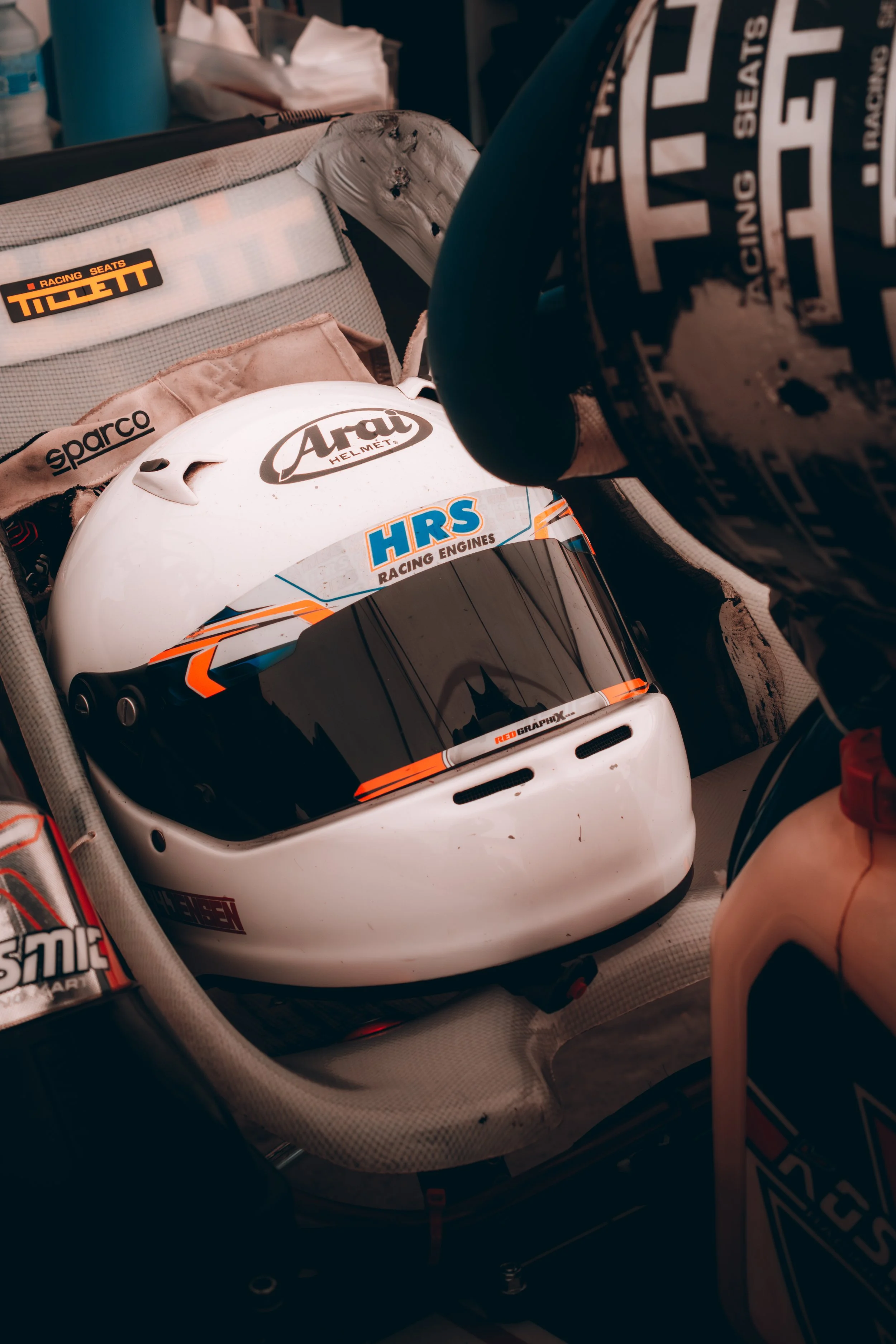 A white racing helmet with various sponsor logos, including 'HRS Racing Engines' and 'Arai,' placed inside a racing vehicle. The helmet is resting on a padded surface with additional racing gear visible nearby.