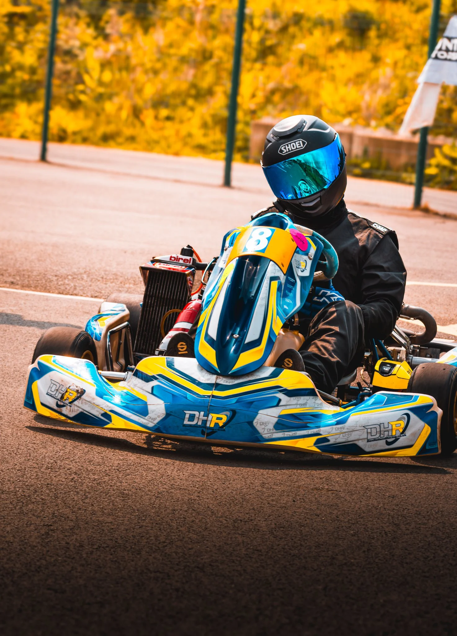 A racer in a black helmet and racing suit sitting on a blue, yellow, and white go-kart with various sponsor logos, on a track during autumn with orange and yellow foliage in the background.